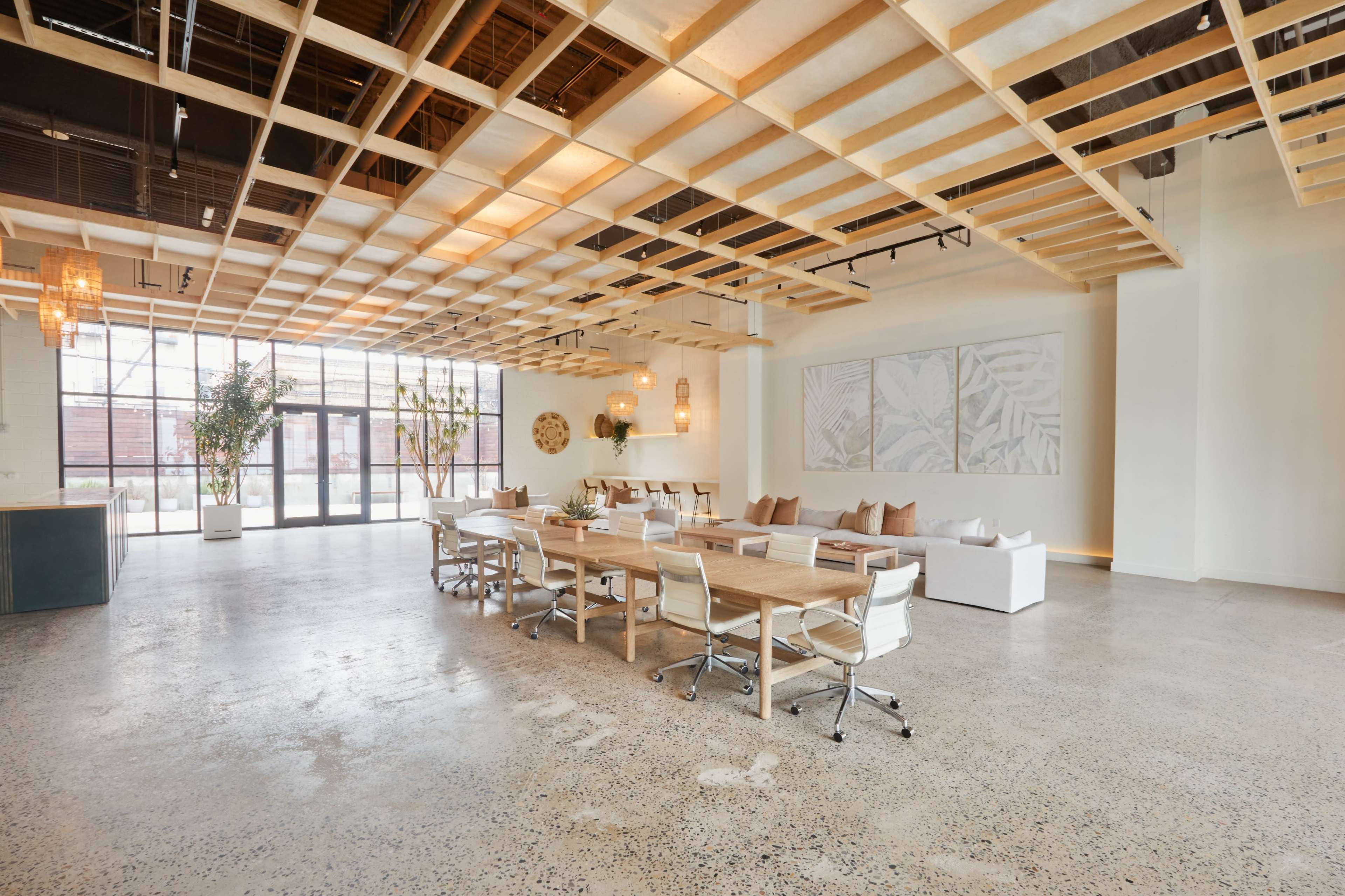 The image shows a spacious and modern office interior featuring a large wooden table surrounded by white chairs, with a wall of windows letting in natural light.