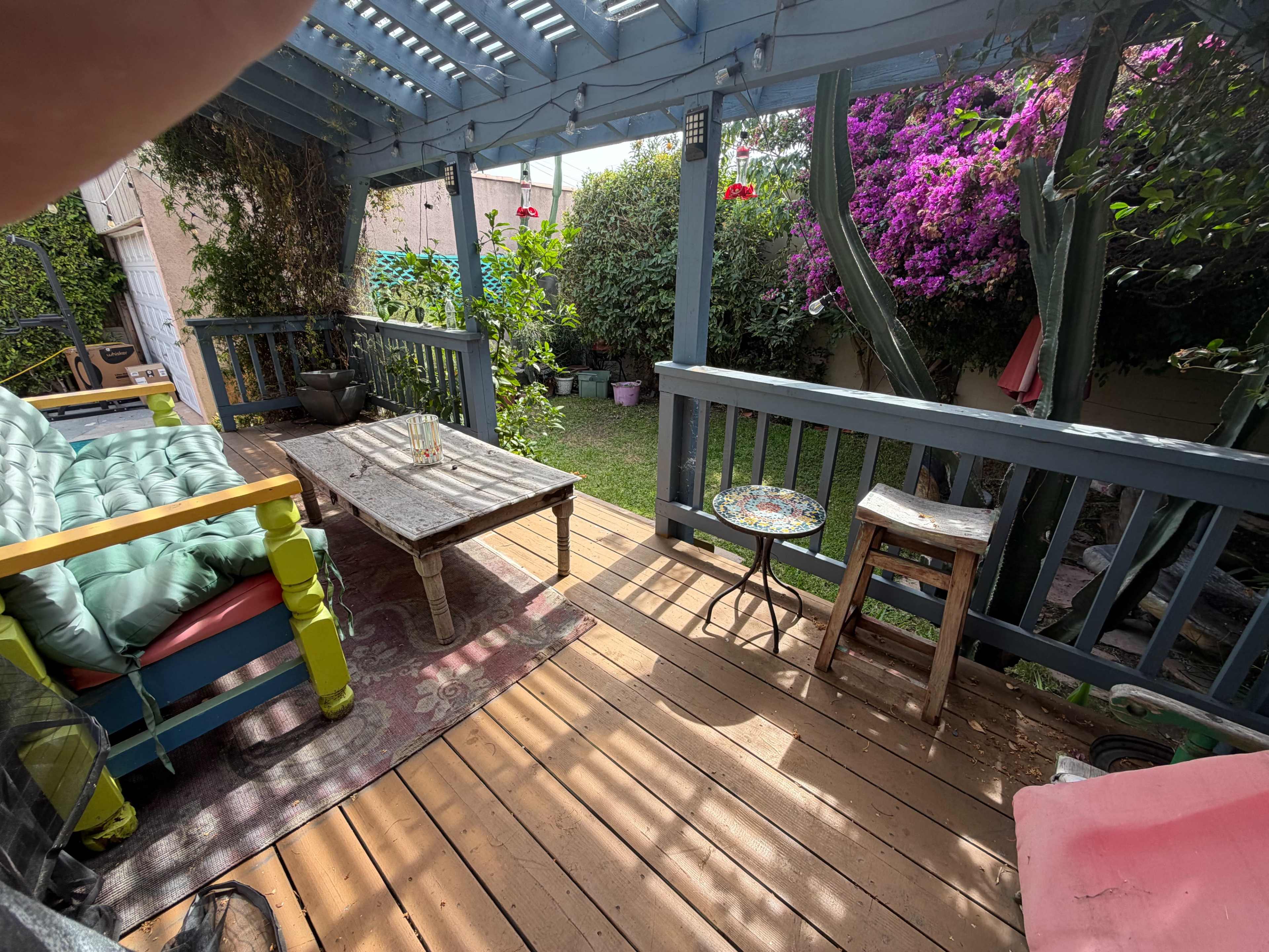 The image shows a wooden deck area with colorful furniture, including a couch and a small table, surrounded by greenery and flowering plants.
