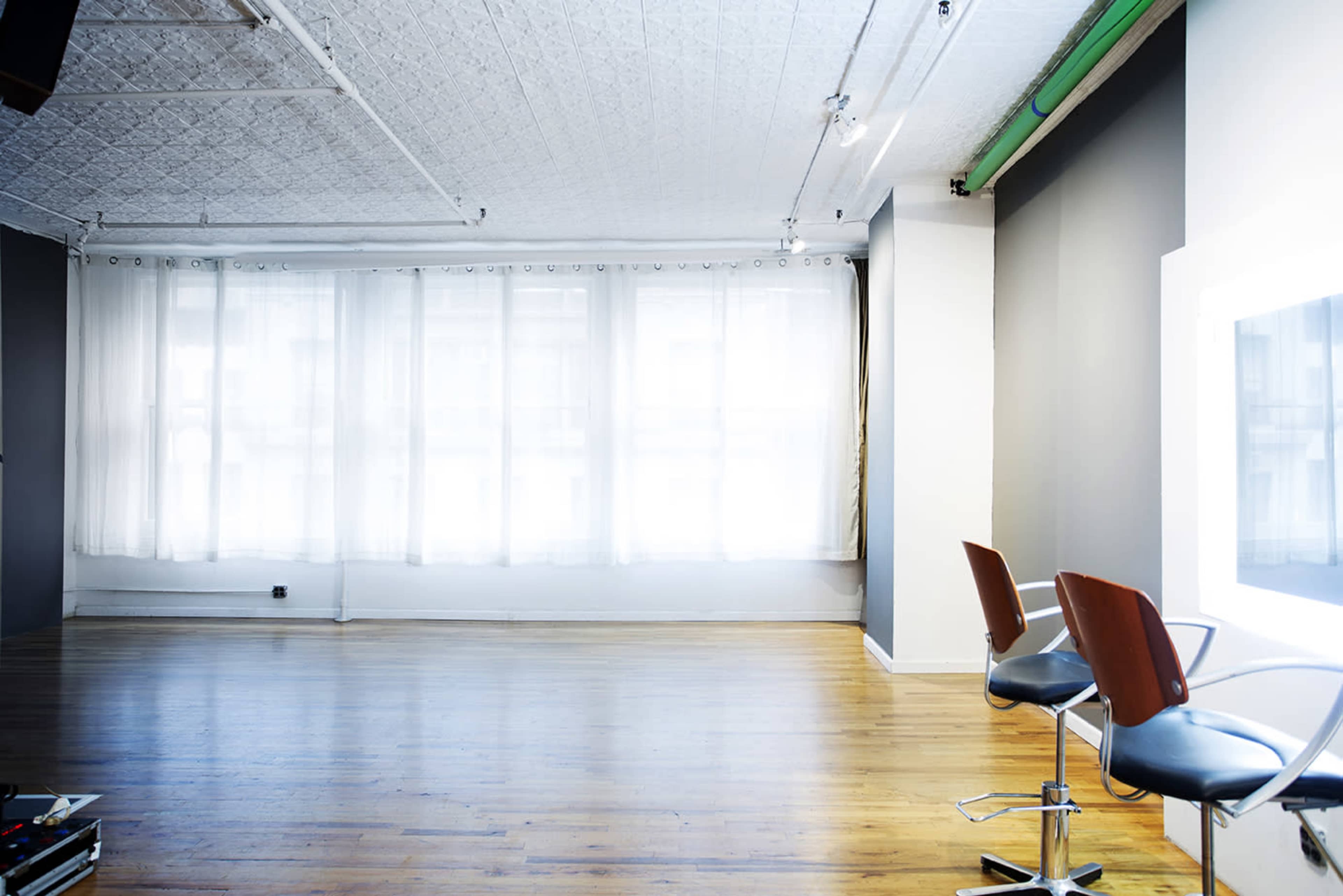 The image shows a bright, empty studio space with large windows, wooden flooring, and two chairs positioned near a lighted mirror.