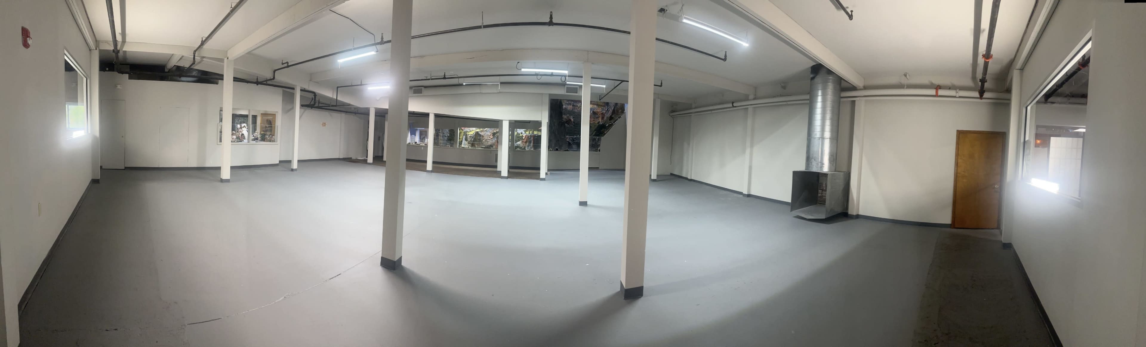 The image shows an empty industrial space with concrete flooring, white walls, and large windows along one side.