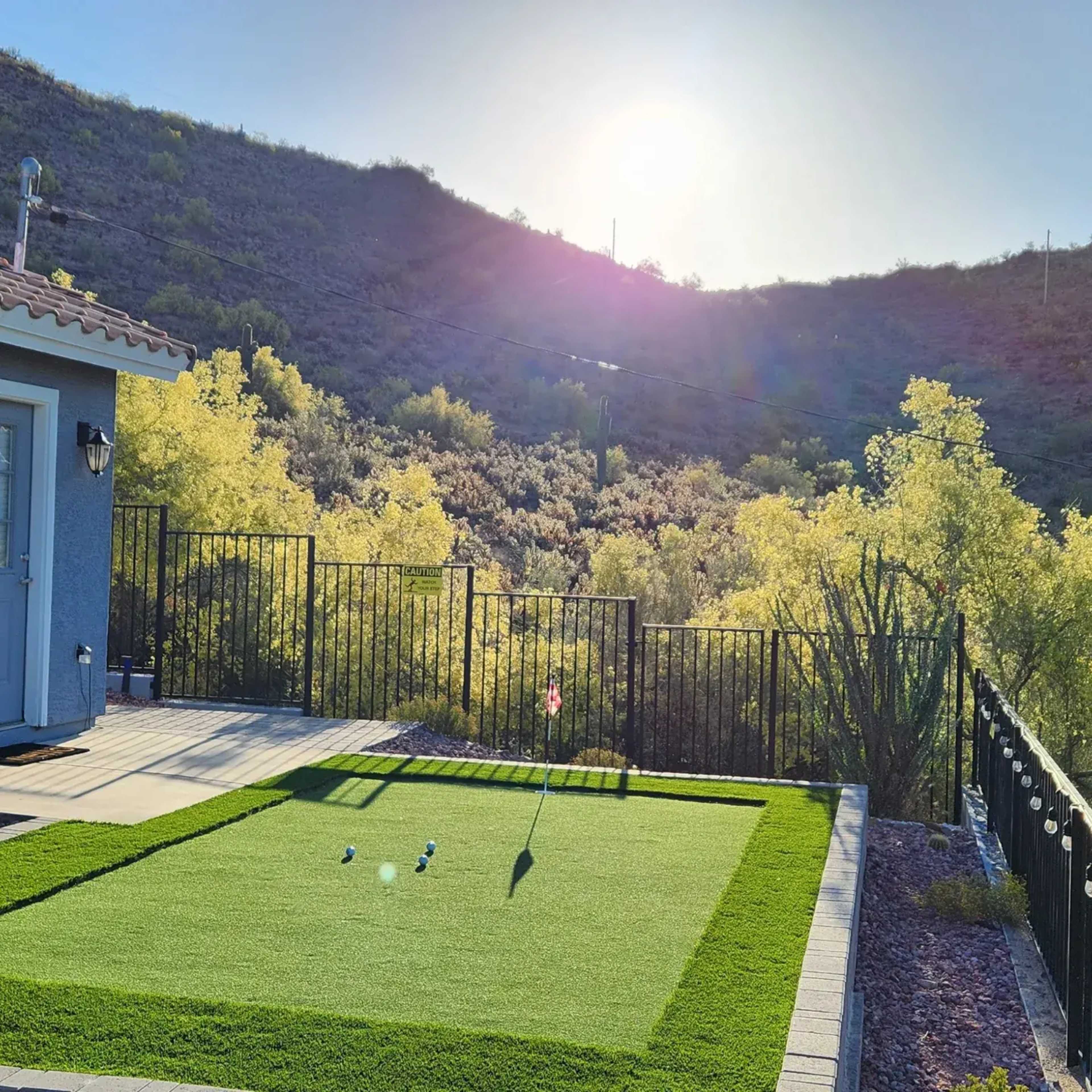 A putting green with two golf balls and a flag is situated in a backyard overlooking a desert hillside under a bright sun.