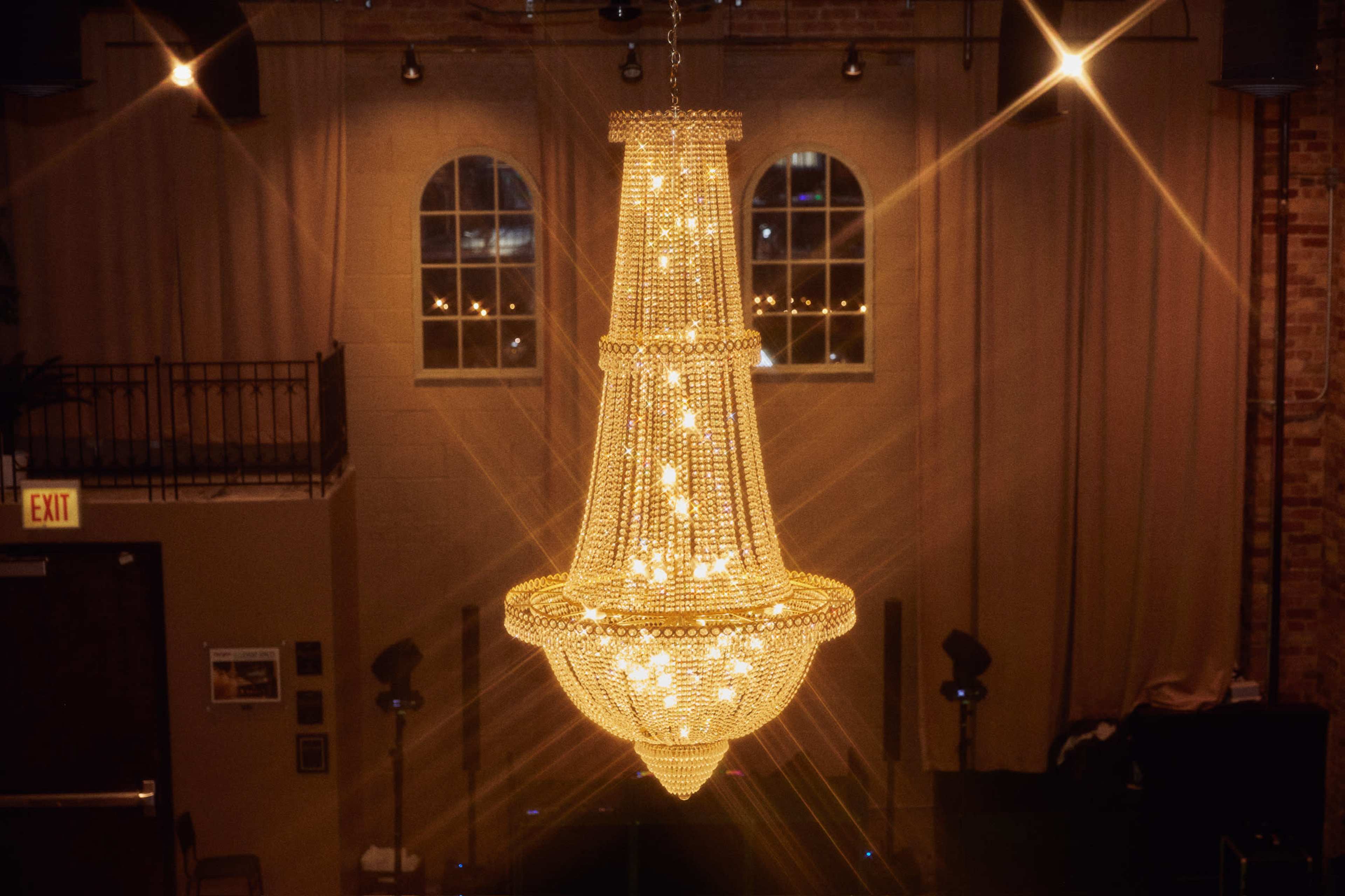 A large, ornate chandelier hangs from the ceiling, illuminated by warm lights, in a room with exposed brick and tall windows.