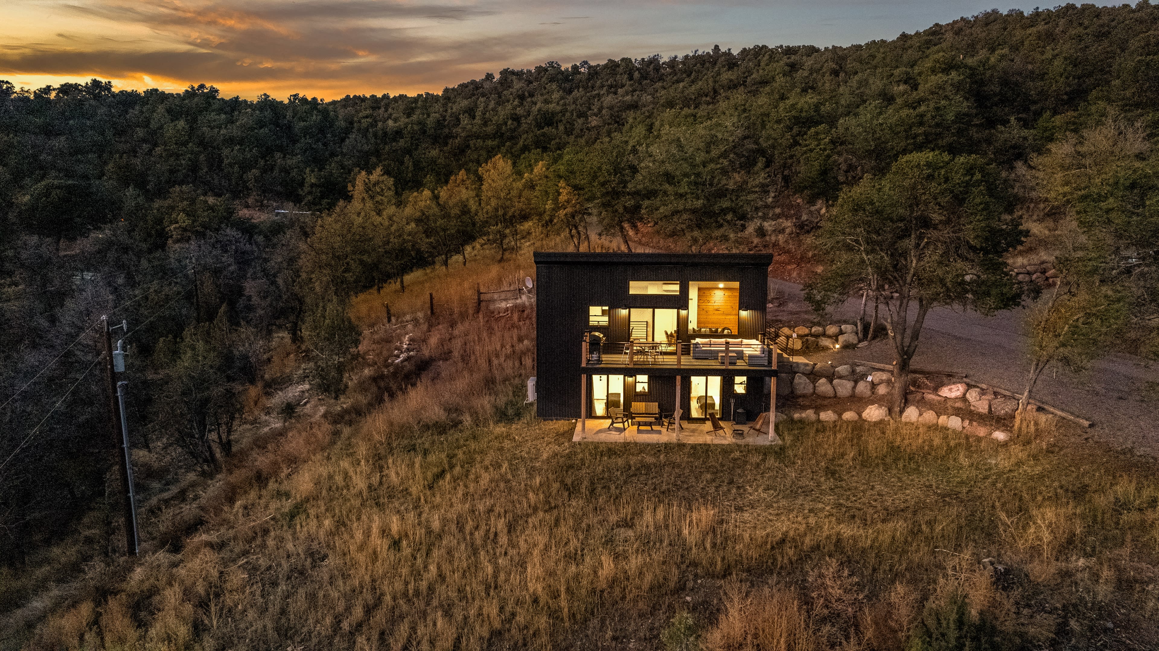 A modern black house sits on a hillside surrounded by trees and rocky terrain at sunset.