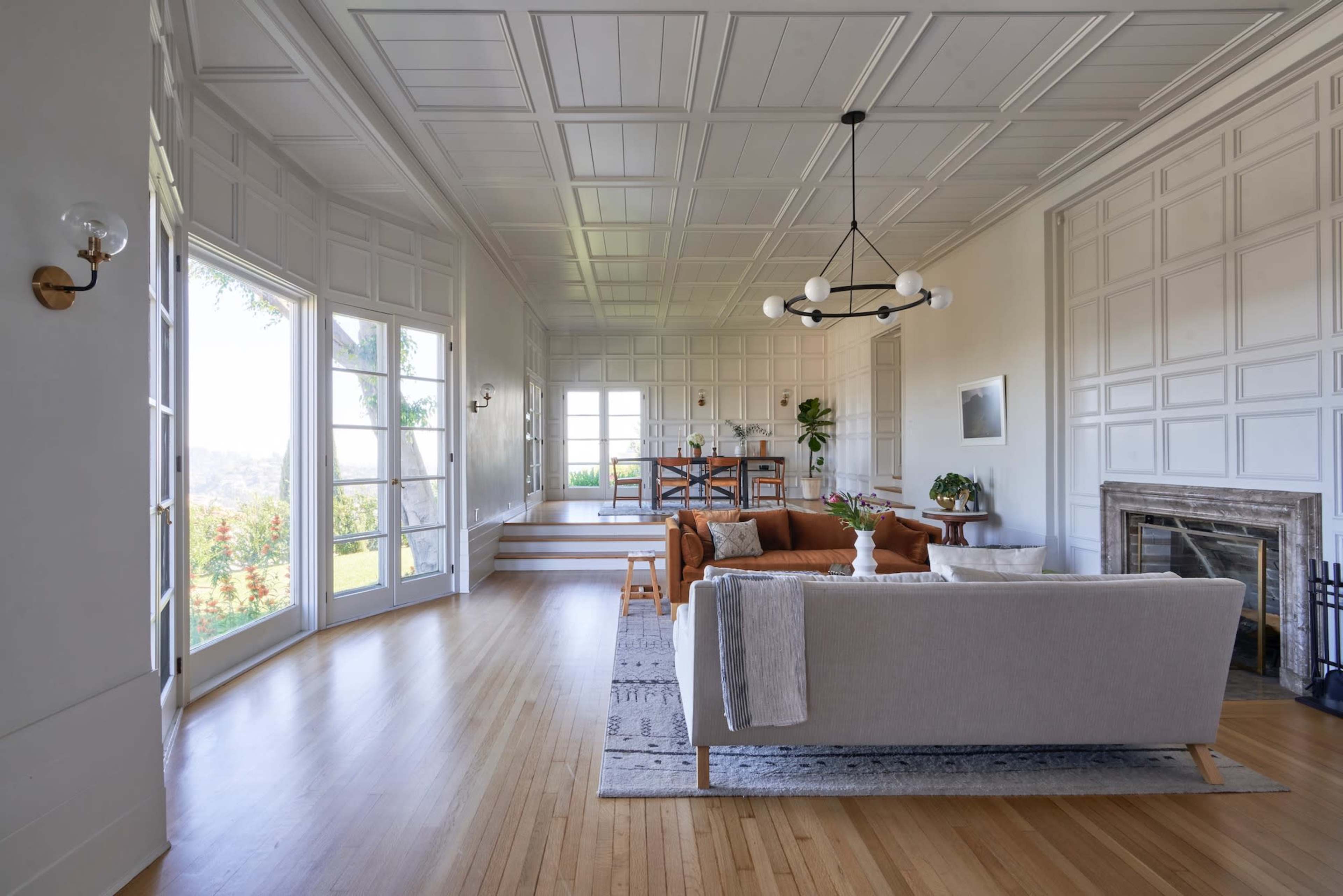 A spacious living room features a light-colored sofa and a brown armchair, with large windows letting in natural light and showcasing a view of the outdoors.