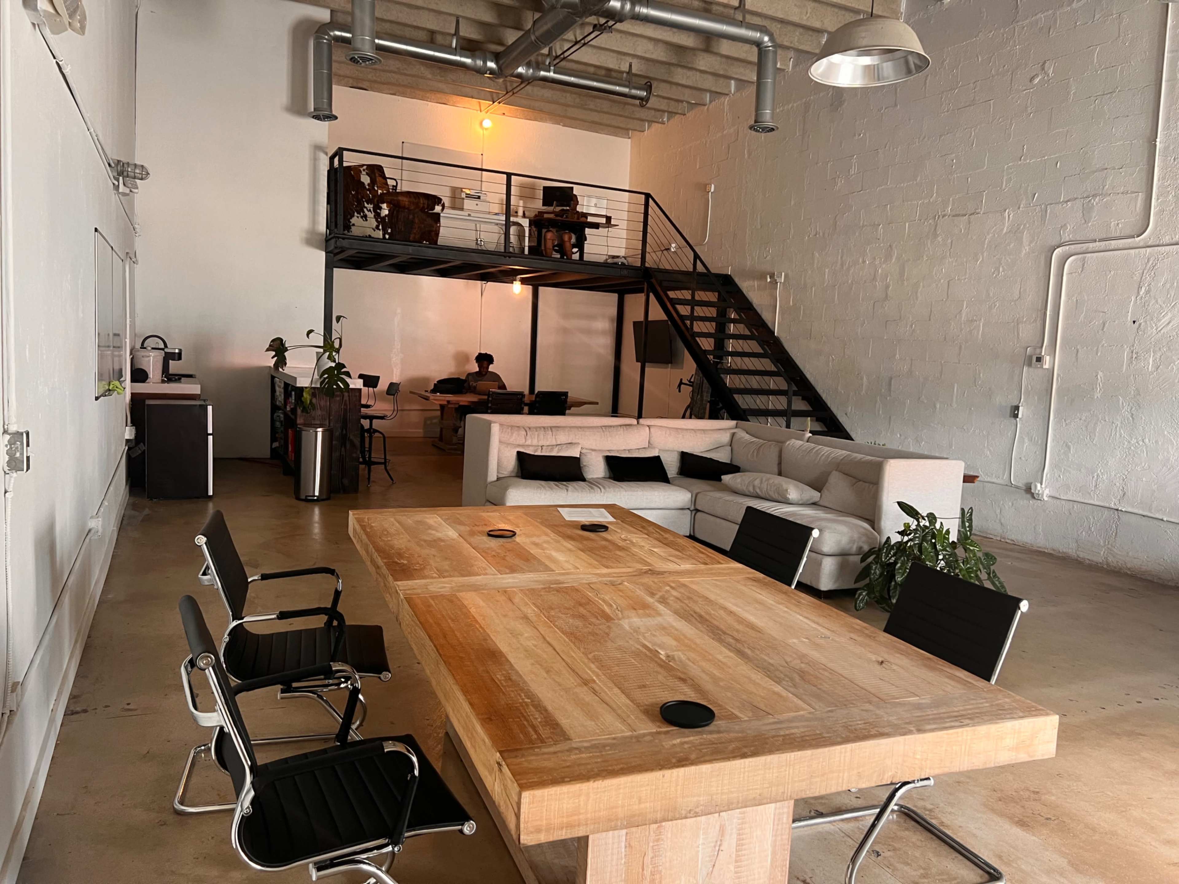A spacious office features a wooden conference table surrounded by black chairs, with a lofted seating area and a couch in the background.