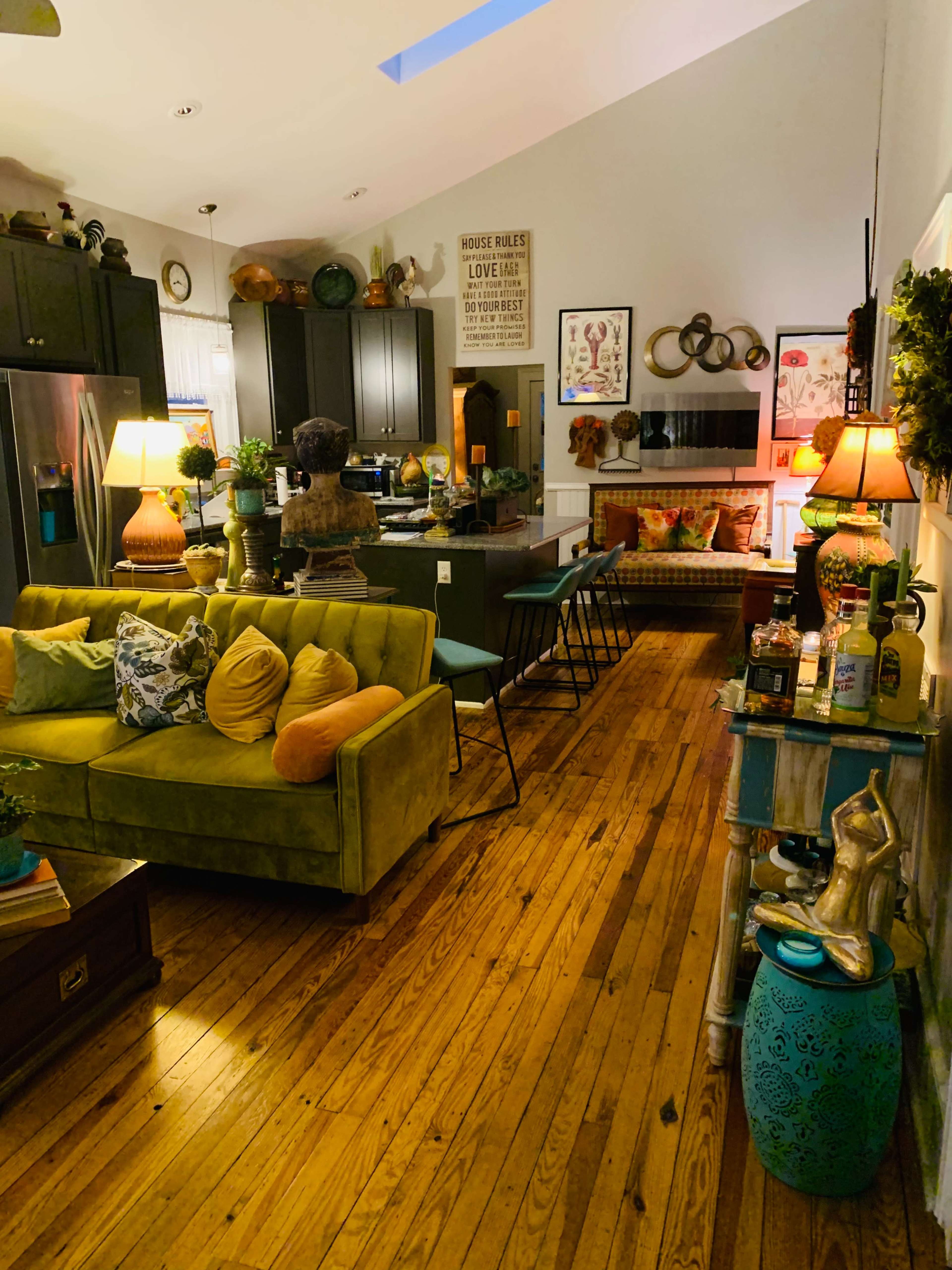A cozy living space with a green couch, wooden floors, and a kitchen area in the background, featuring various decorative elements and furnishings.