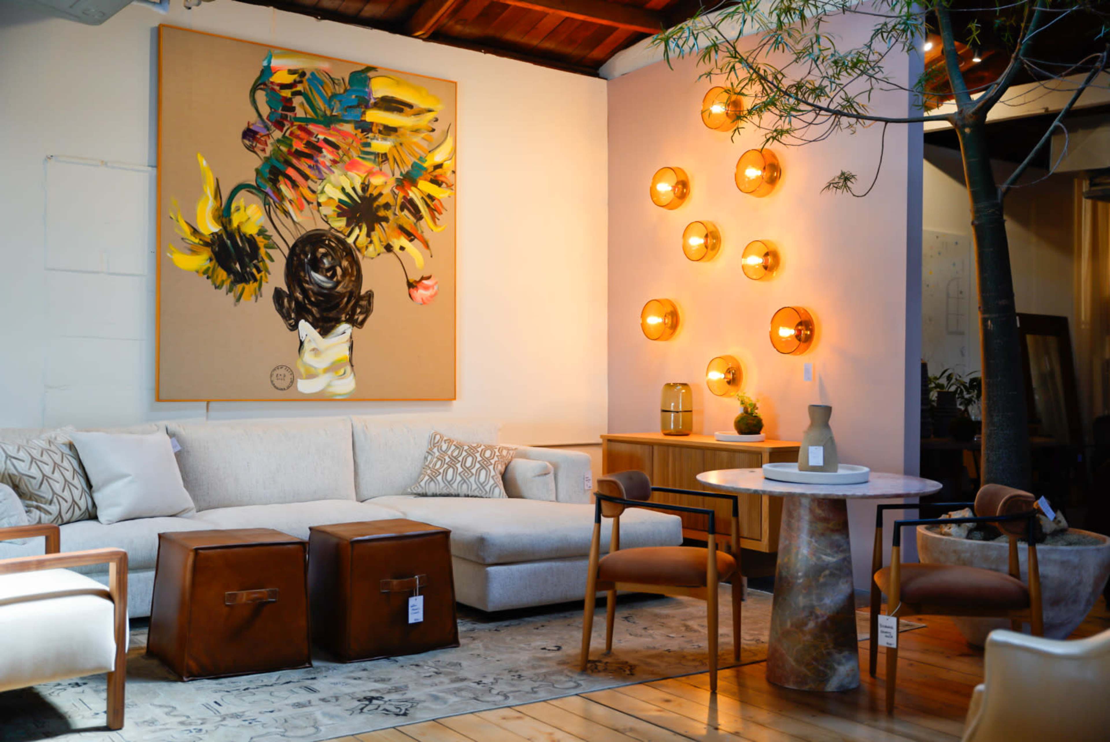 The image showcases a modern living room with a beige sectional sofa, a round marble table, and a large colorful artwork on the wall, complemented by decorative lighting fixtures.