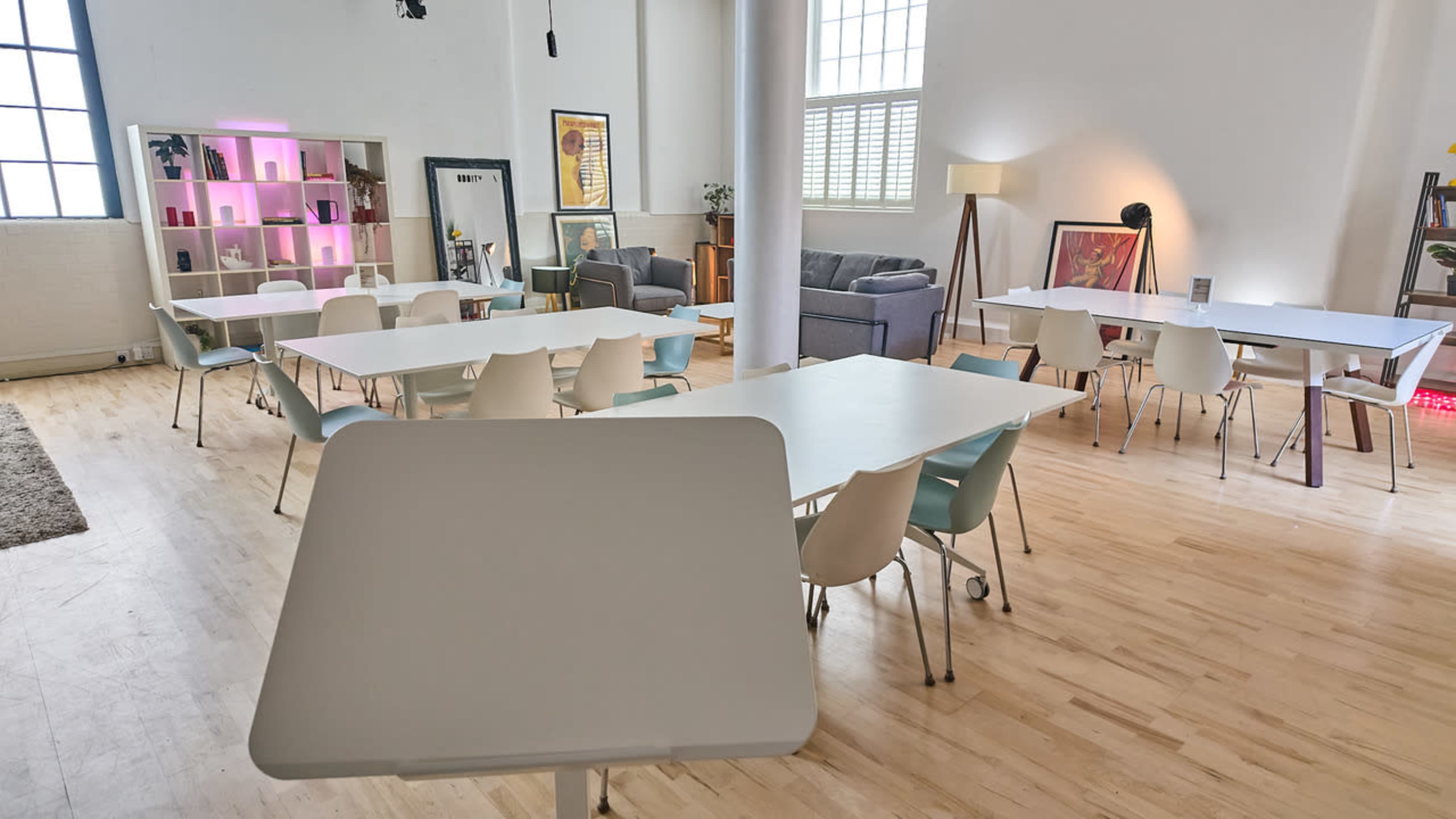 The image shows a spacious, brightly lit room filled with several tables and chairs, with a seating area featuring couches and decorative artwork.