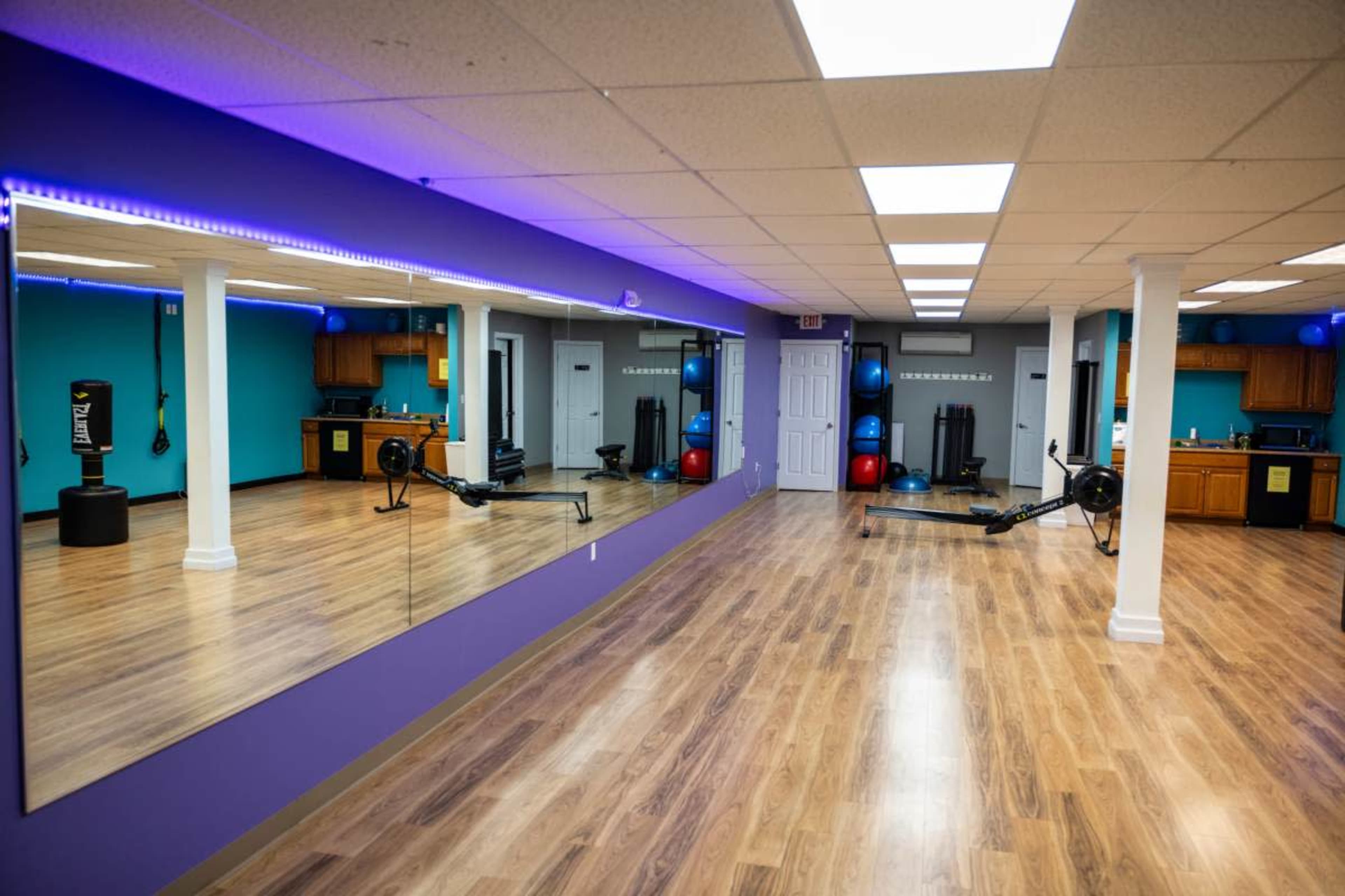 A fitness studio features wooden flooring, blue walls, and exercise equipment, including an rowing machine and stability balls.