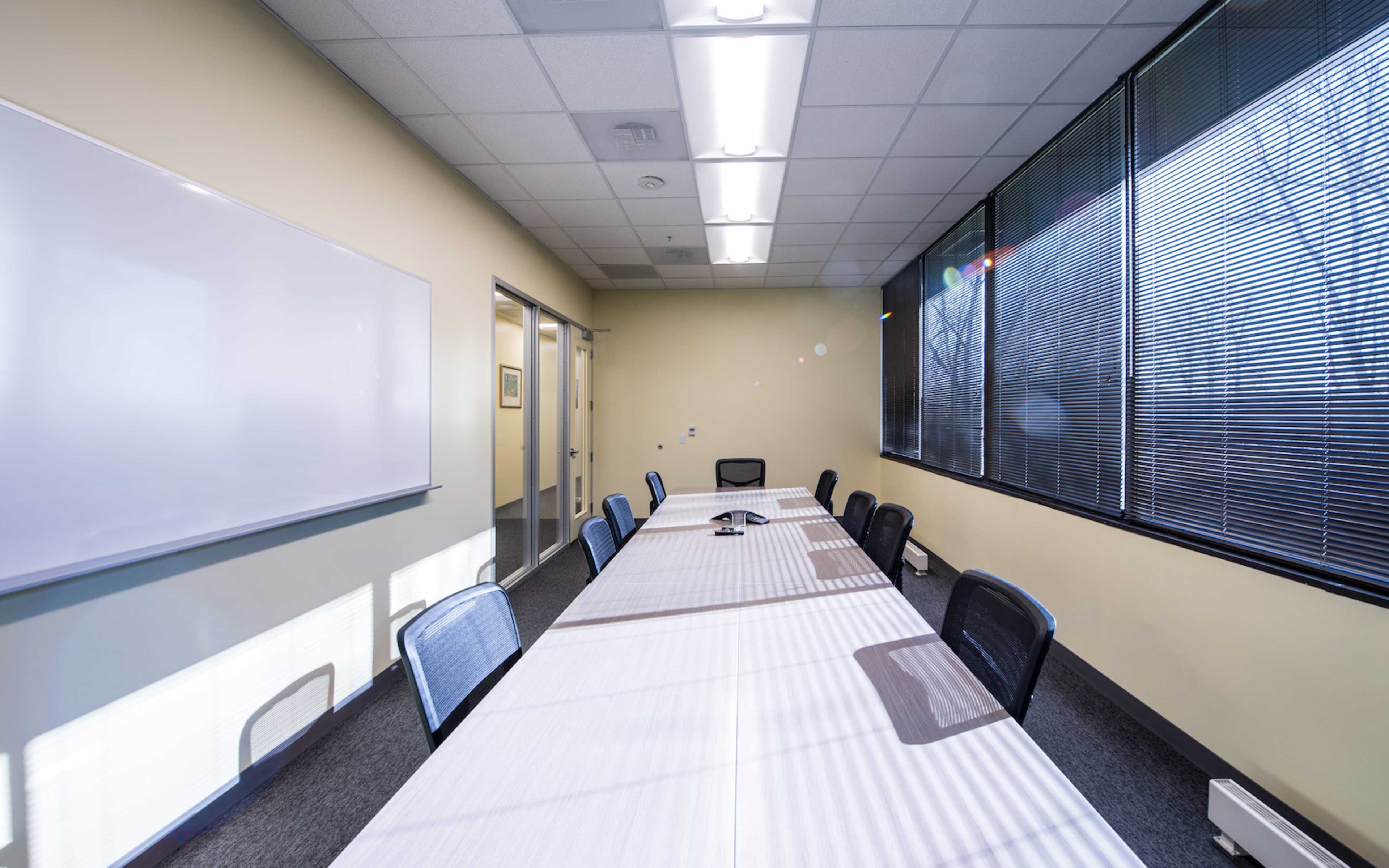 A long meeting table with several black chairs is set up in a brightly lit conference room with large windows.
