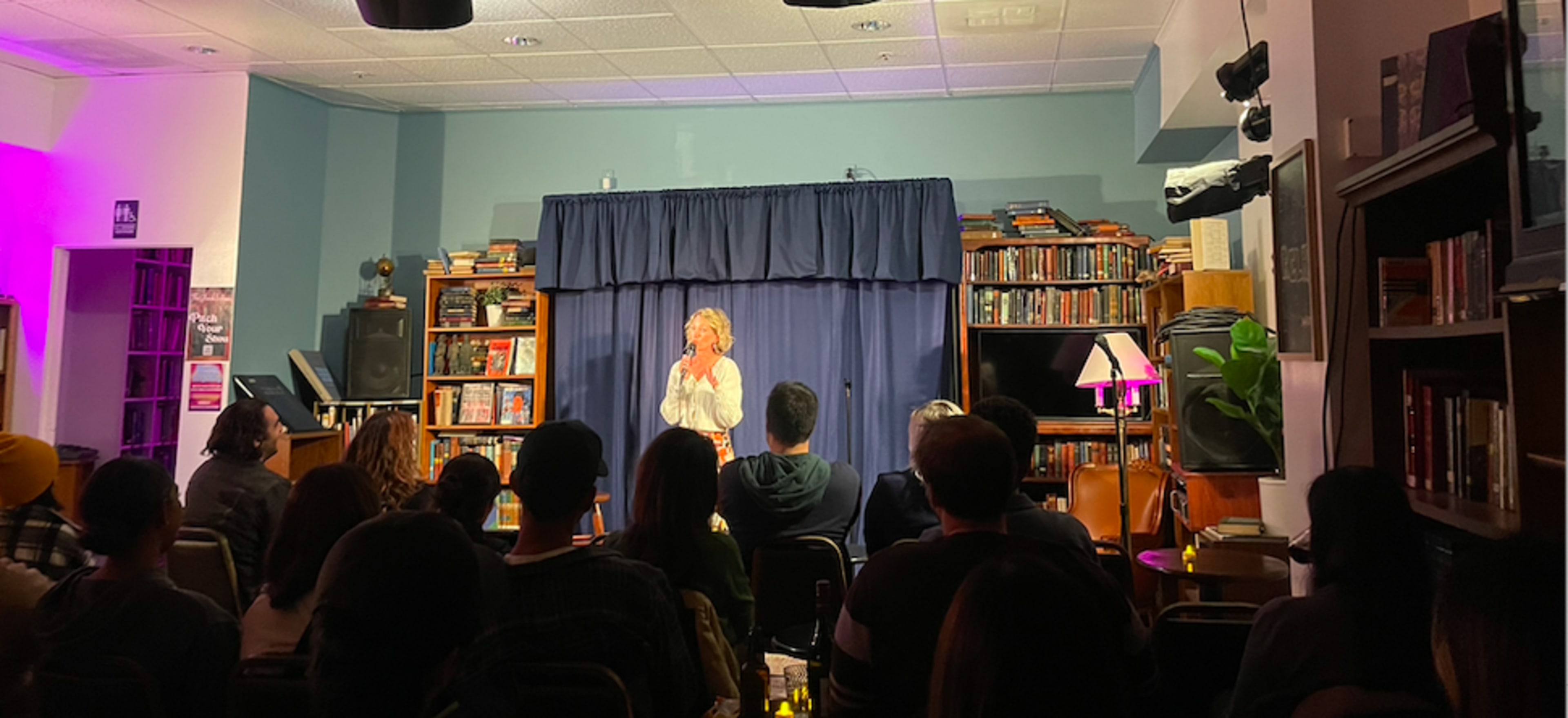 A performer stands on stage in front of an audience, illuminated by a spotlight, with bookshelves and lamps in the background.