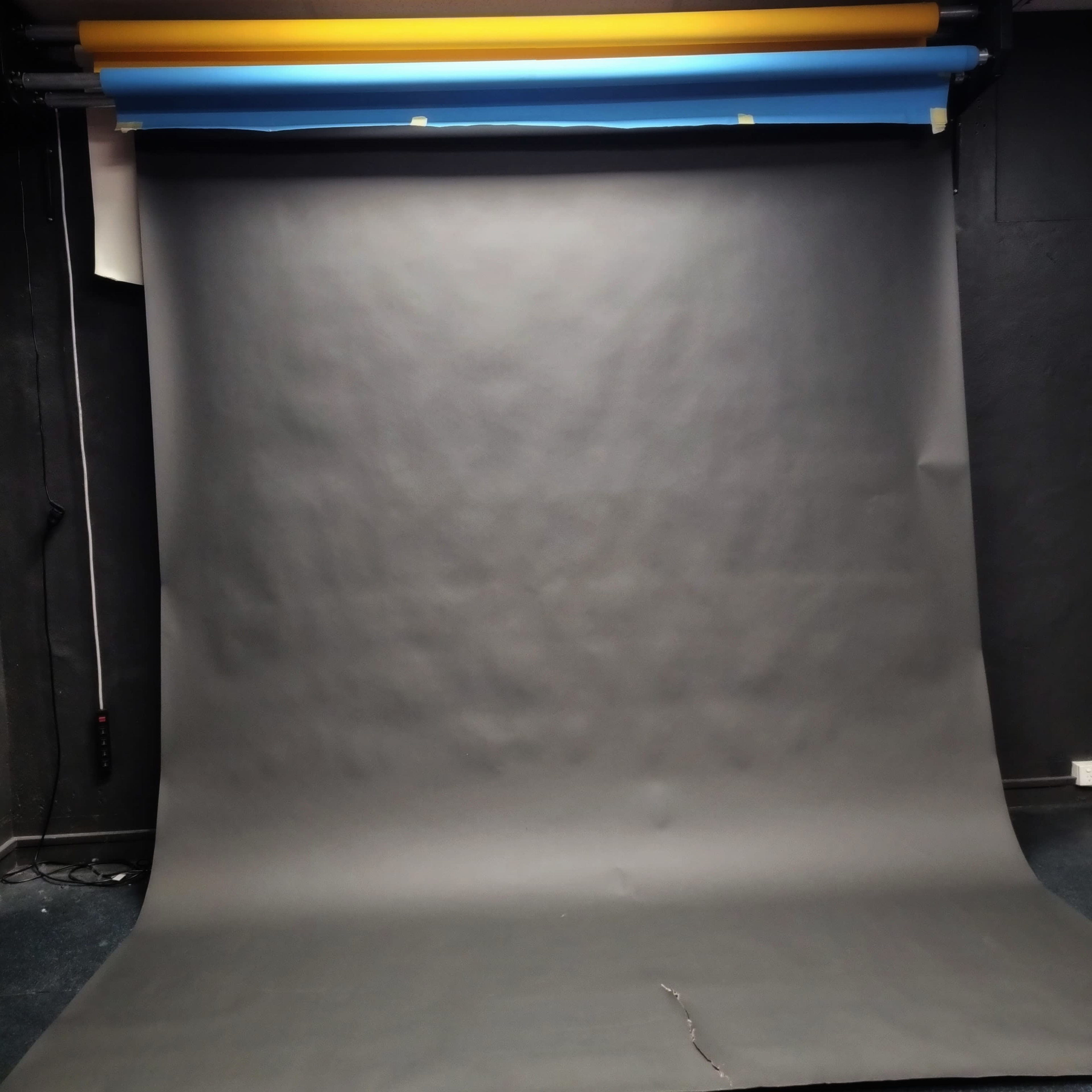 A photography backdrop made of gray fabric, with rolls of blue and yellow paper hanging above it.