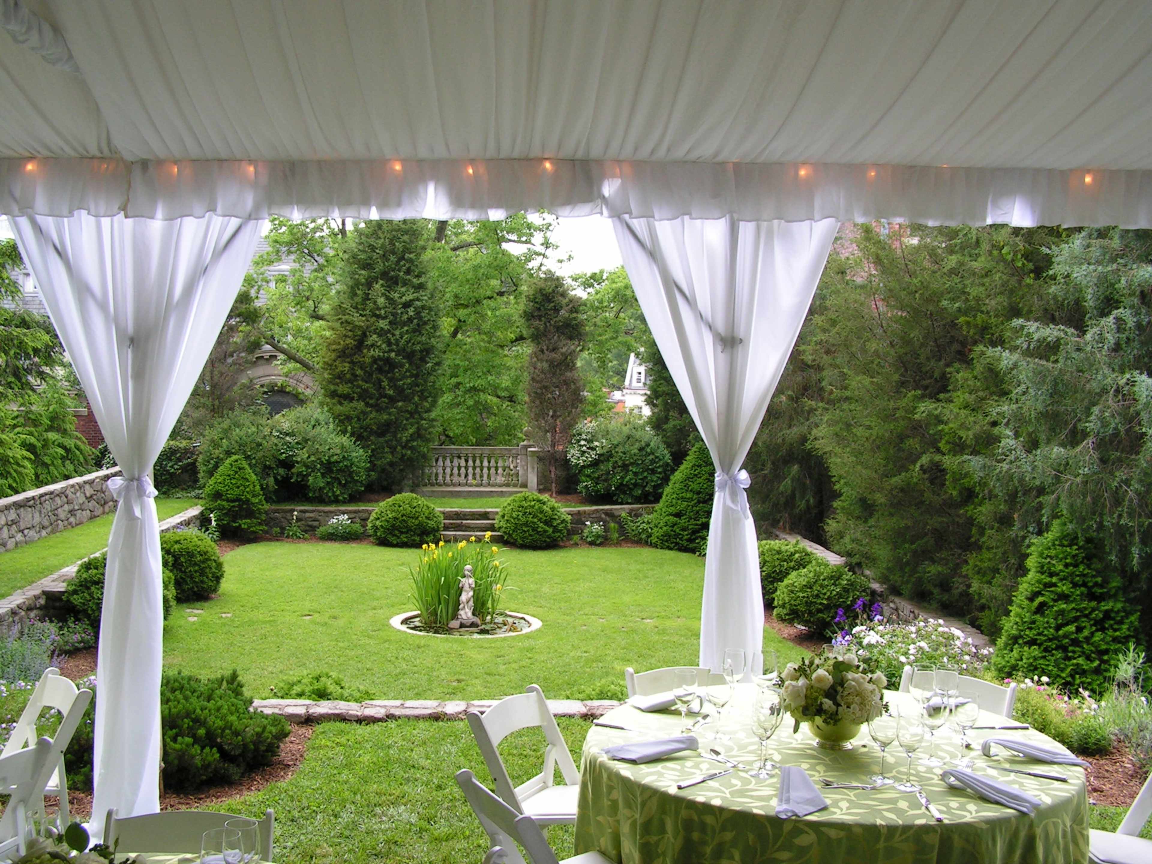 A tented outdoor setting with a round dining table set for a meal, overlooking a lush garden with neatly trimmed shrubs and a central fountain.
