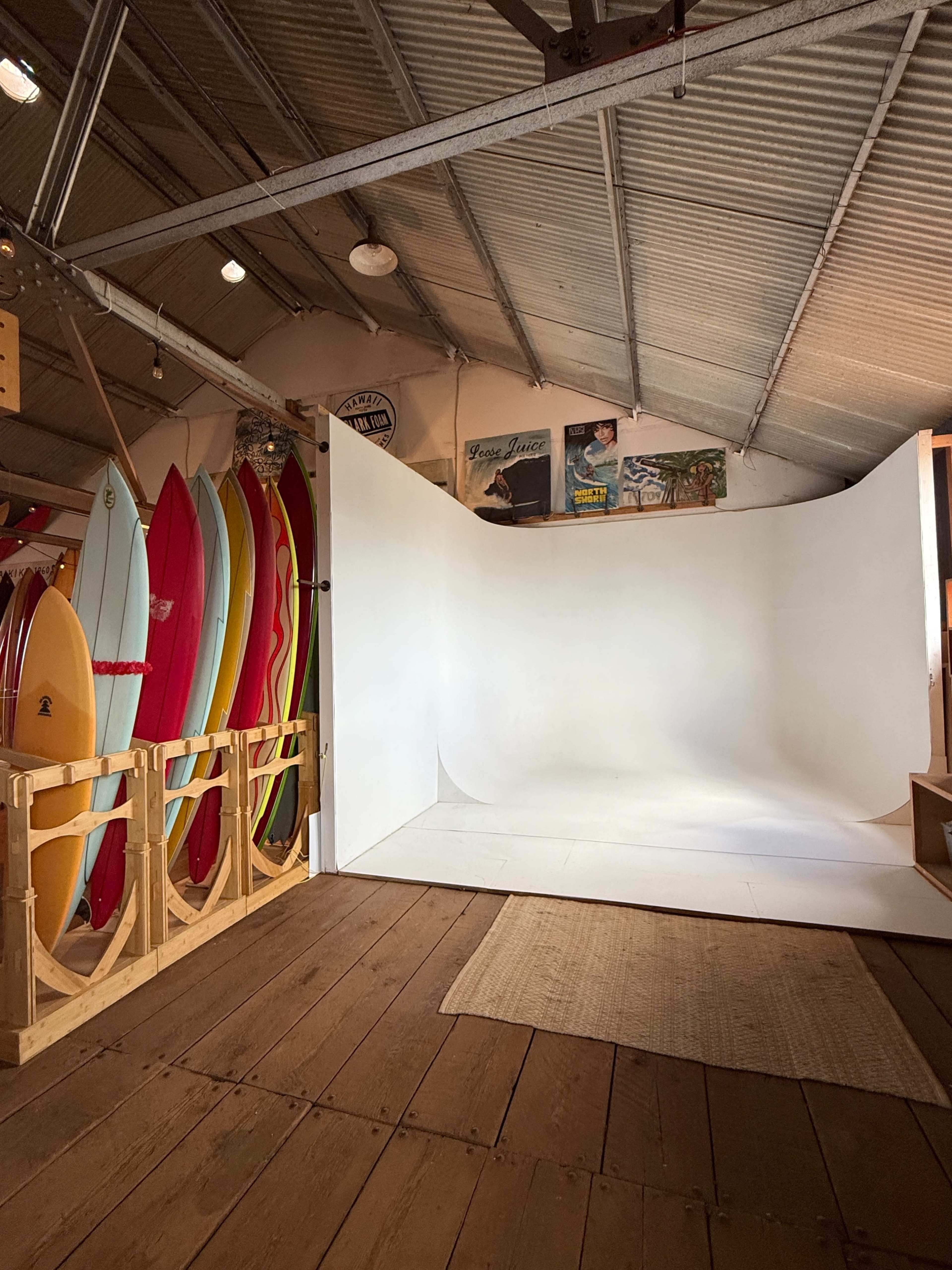 The image shows a spacious indoor area with a white backdrop intended for photography, surrounded by colorful surfboards leaning against the wall.