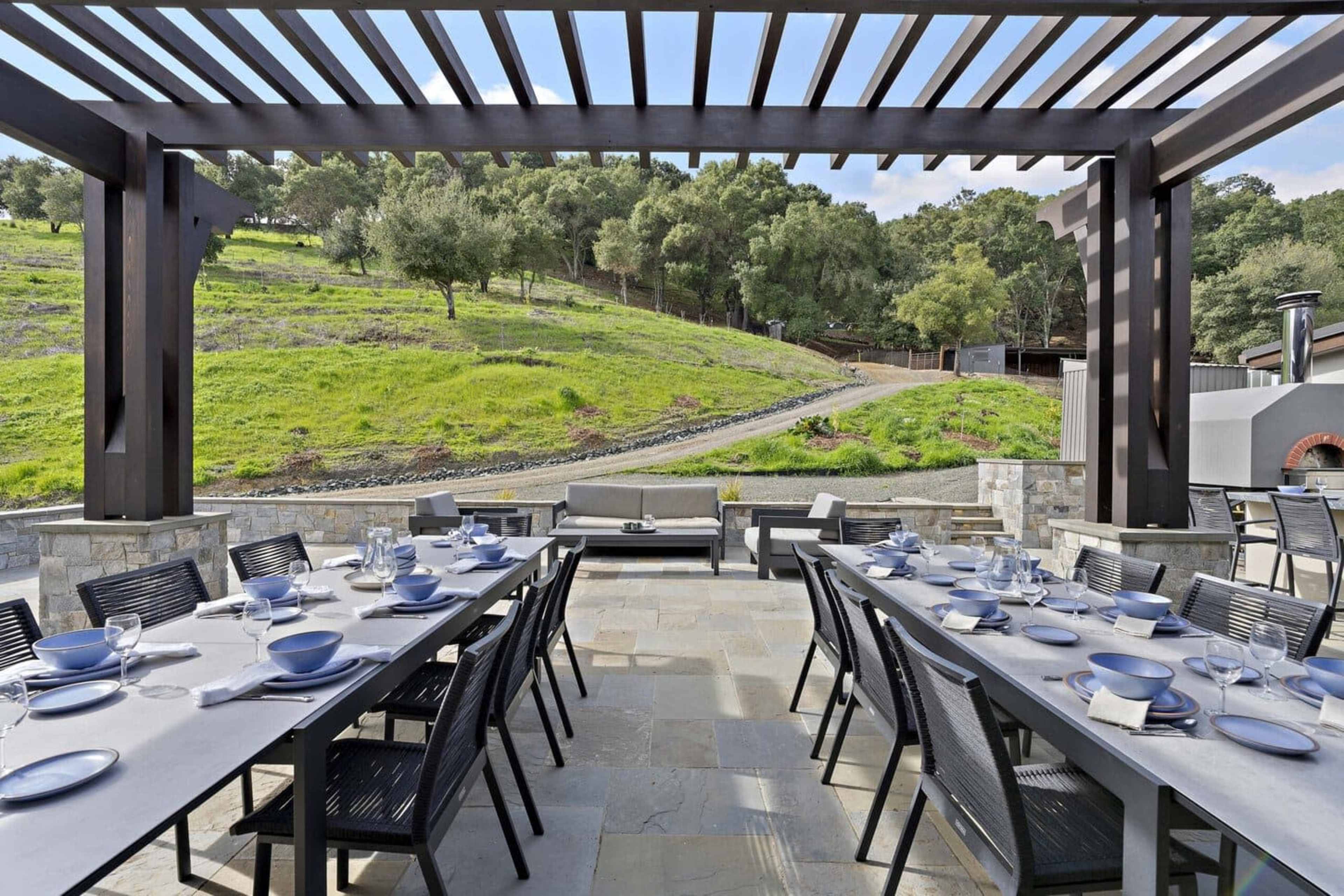 The image shows a spacious outdoor dining area with a long table set for a meal, surrounded by a grassy landscape and shaded by a wooden pergola.