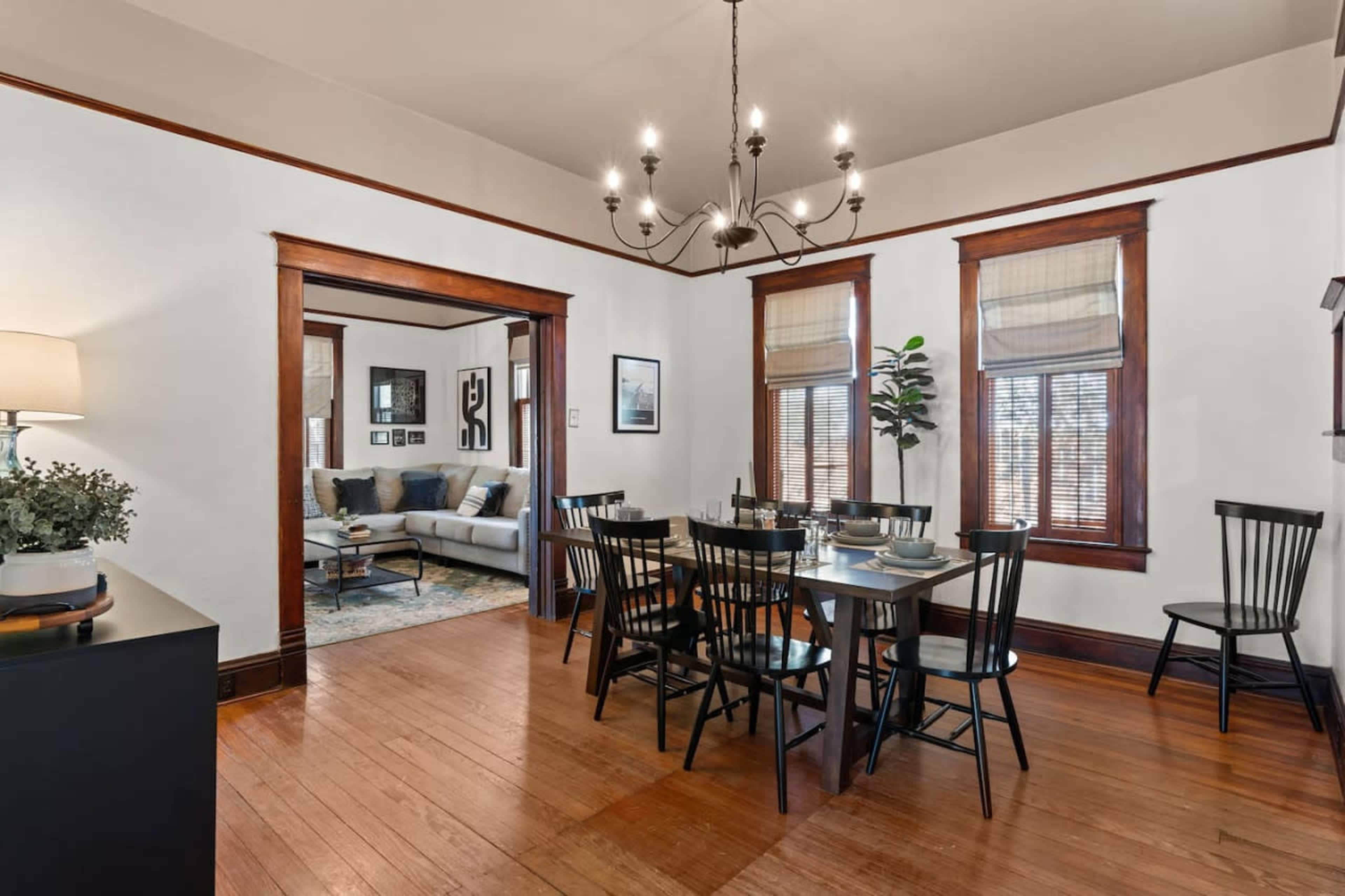 A dining area featuring a table surrounded by black chairs, with a chandelier overhead and a view into a living room through an open doorway.