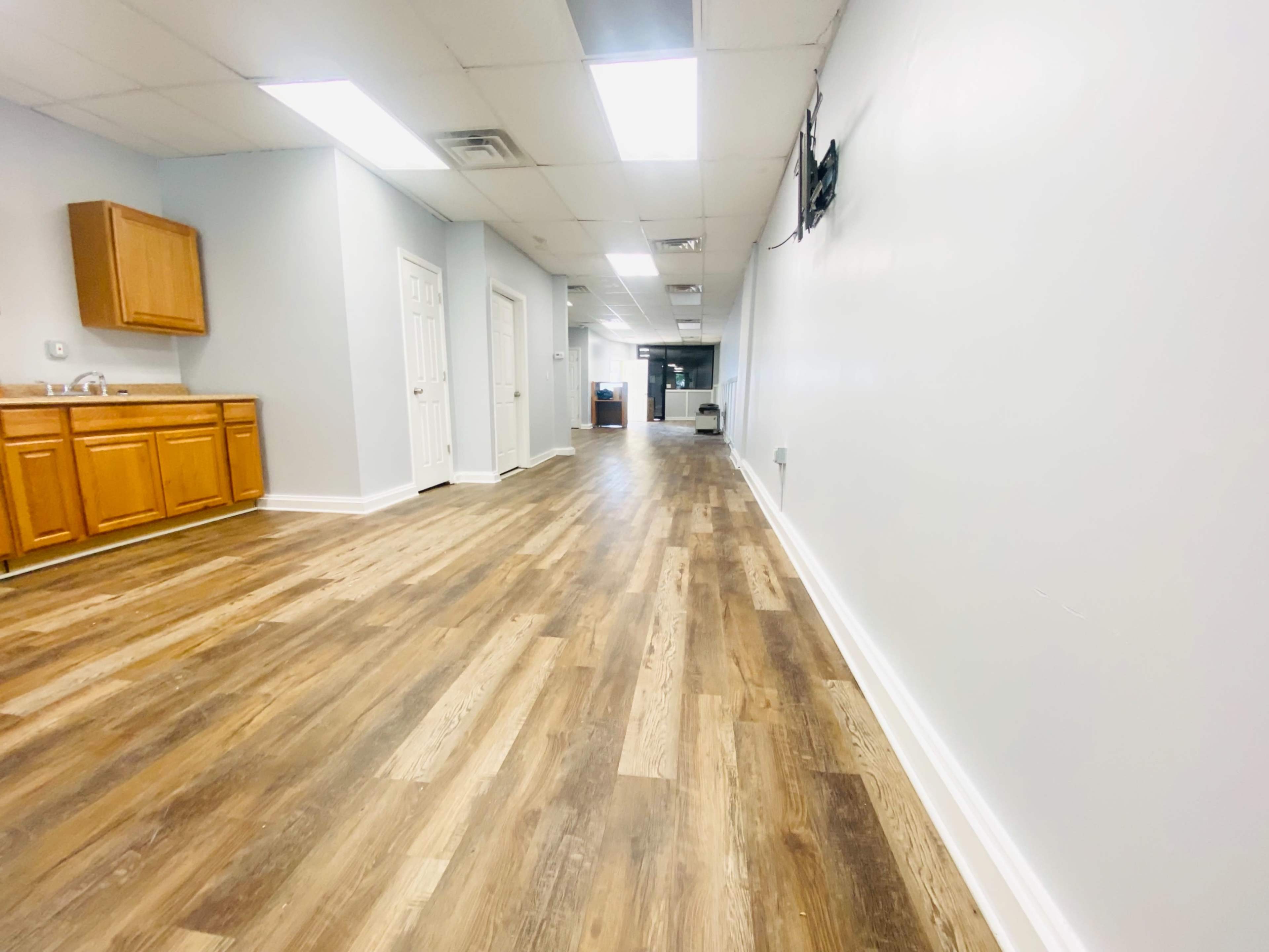 A long hallway with wooden flooring, leading to a waiting area and a kitchenette on the left side.