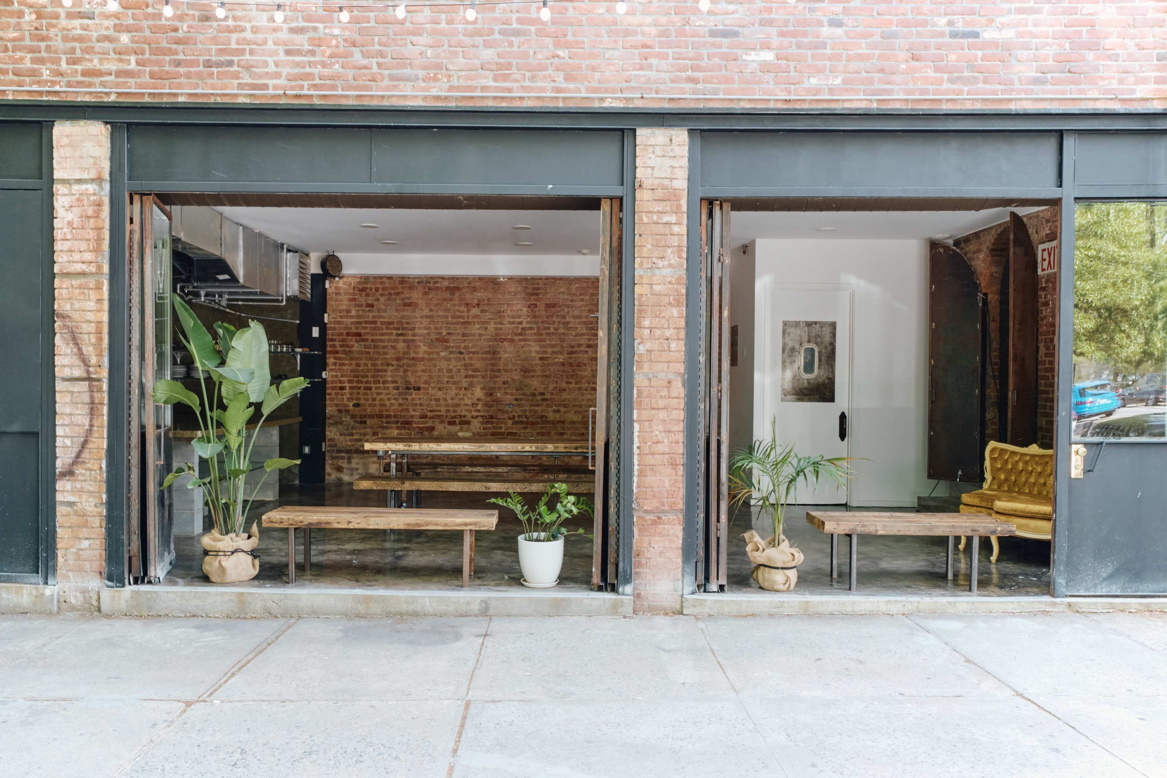 A brick-front building with large windows revealing an interior featuring wooden benches, plants, and a yellow couch.