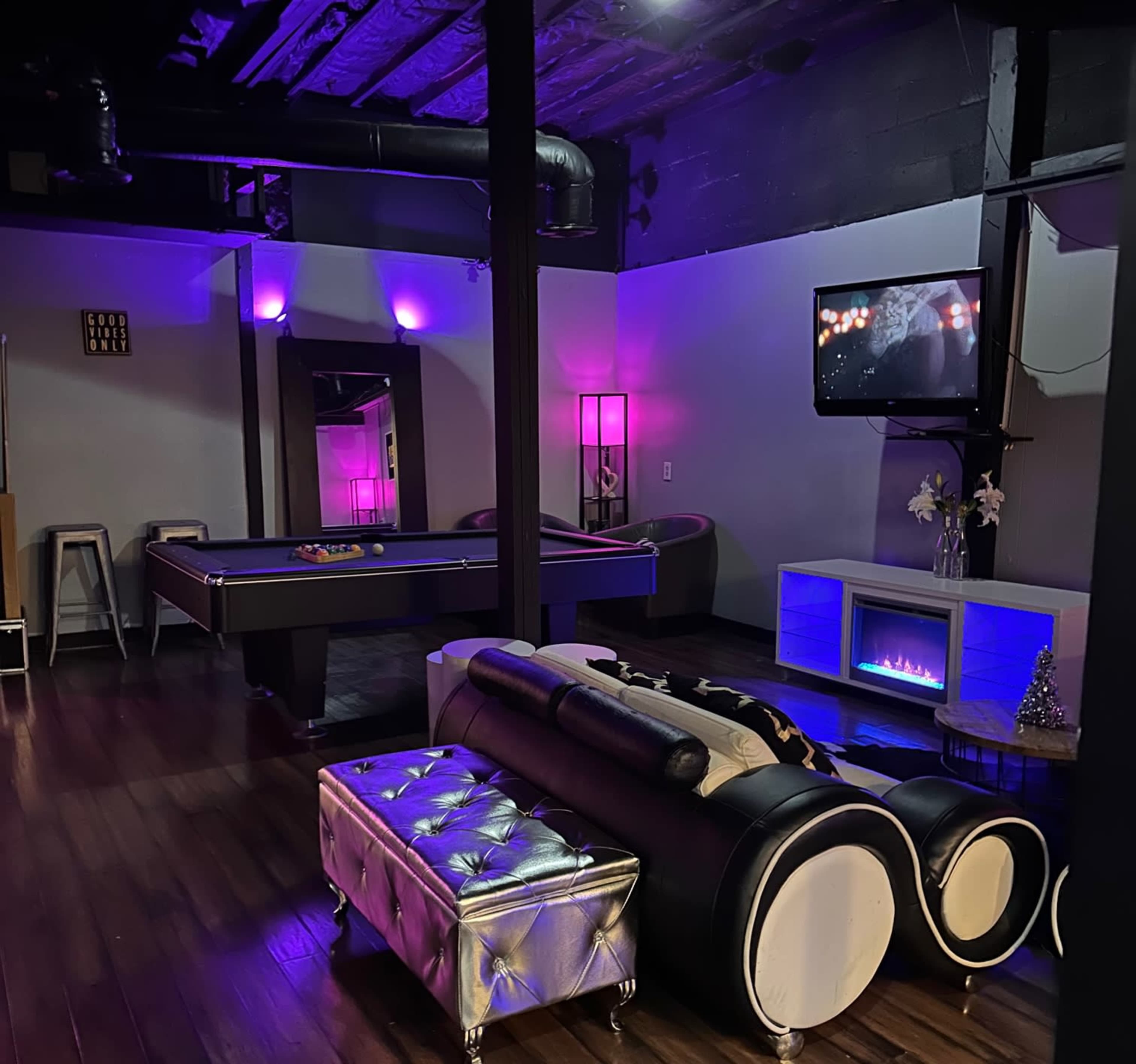 A modern entertainment room featuring a pool table, a black and white sectional sofa, a mounted television, and ambient purple lighting.