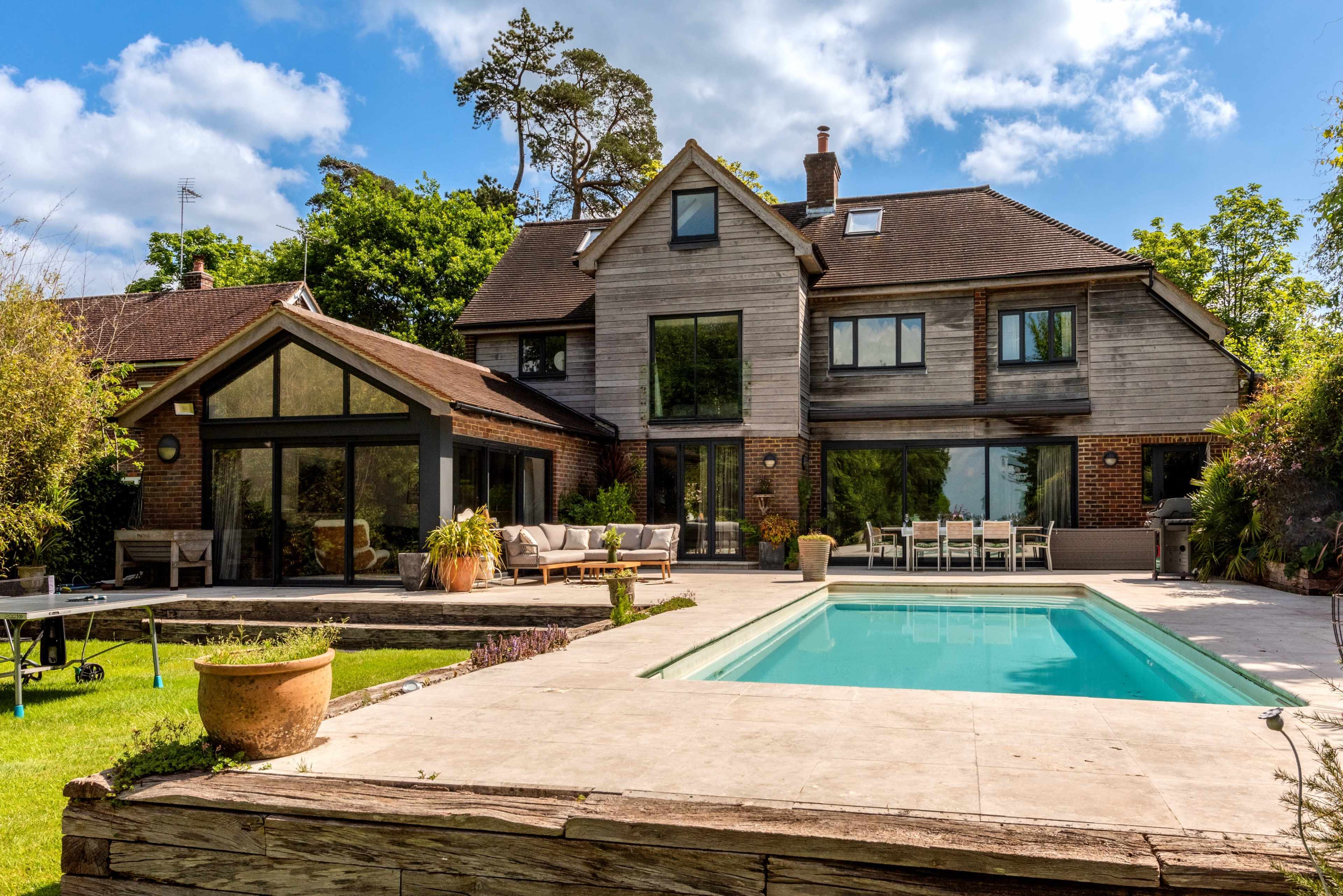 A modern two-story house with large windows overlooks a rectangular swimming pool and a spacious patio, surrounded by greenery.