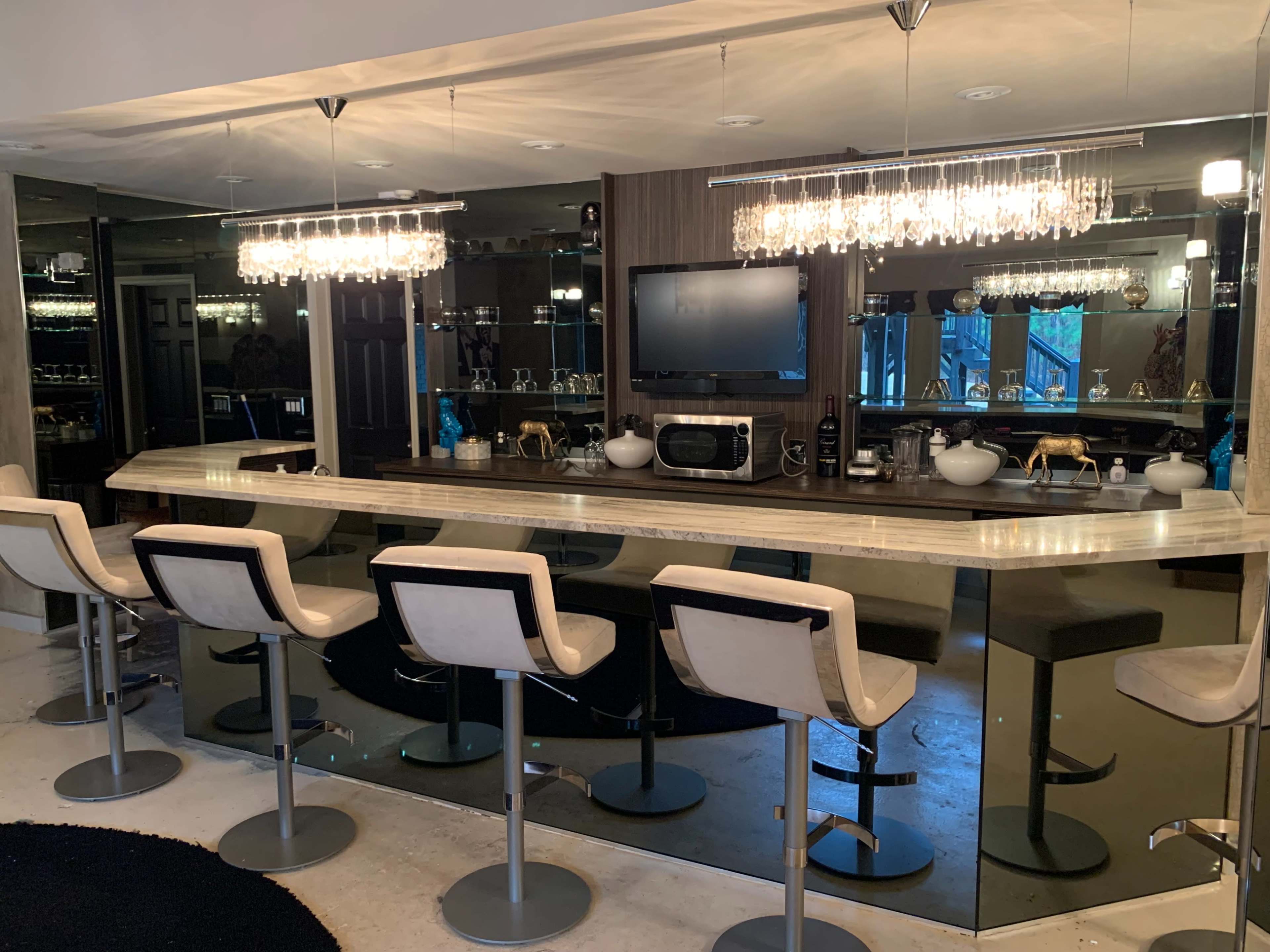 A modern home bar features a polished countertop, high-backed stools, and two sparkling chandeliers above, set against a mirrored wall.