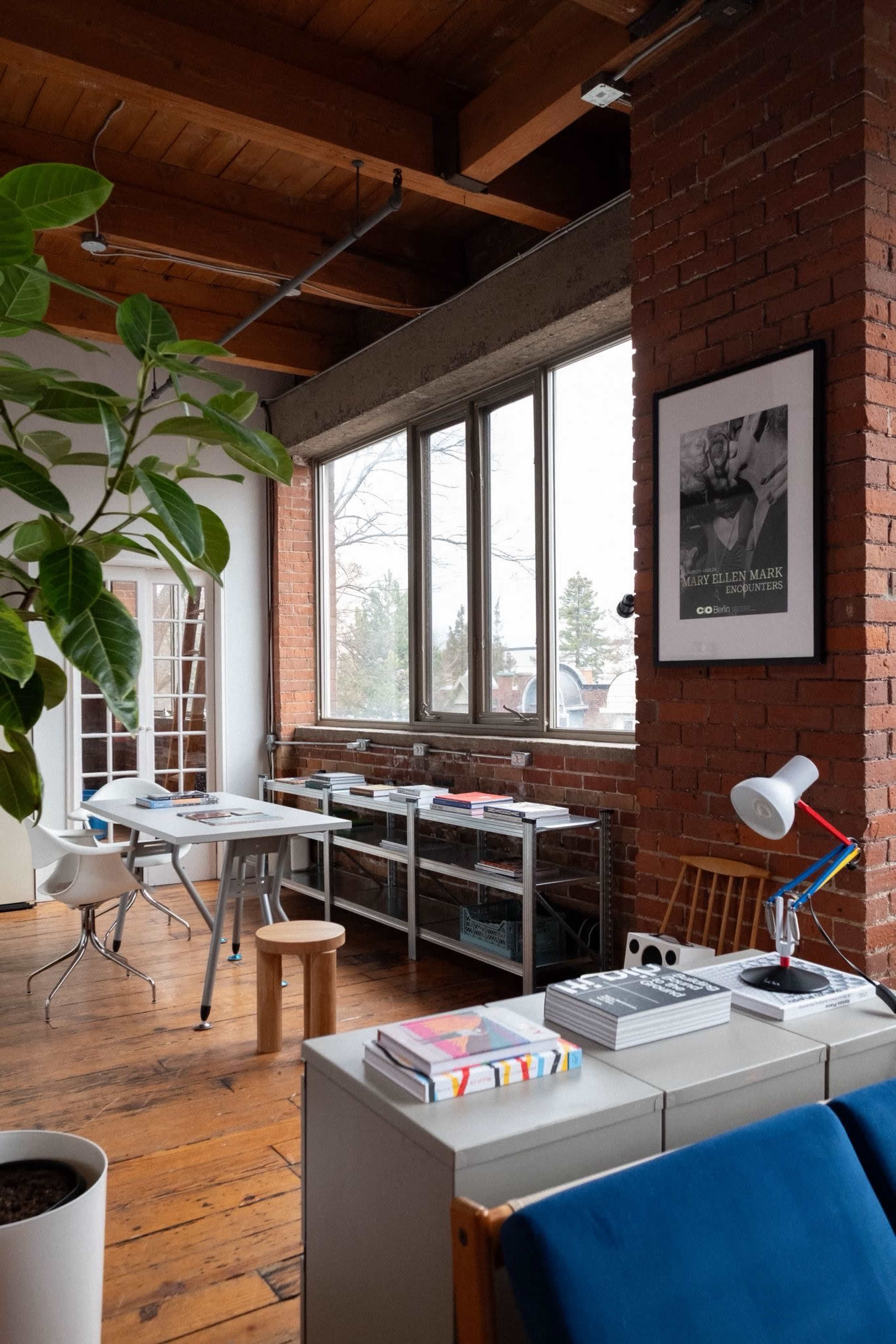 The image shows a bright, airy workspace with large windows, wooden floors, and a mix of modern and vintage furniture.