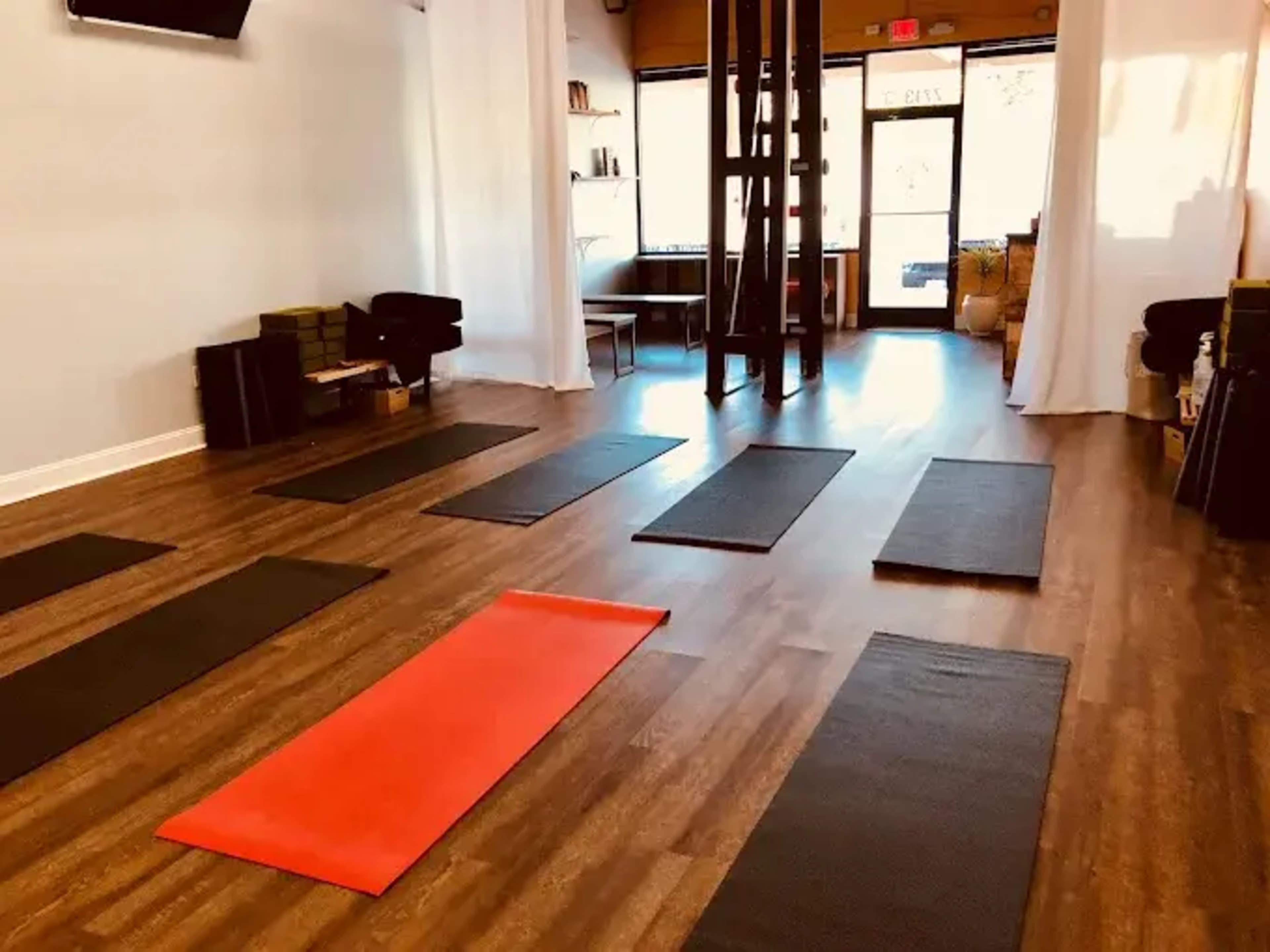 The image shows a yoga studio with several exercise mats arranged on a wooden floor, illuminated by natural light from large front windows.