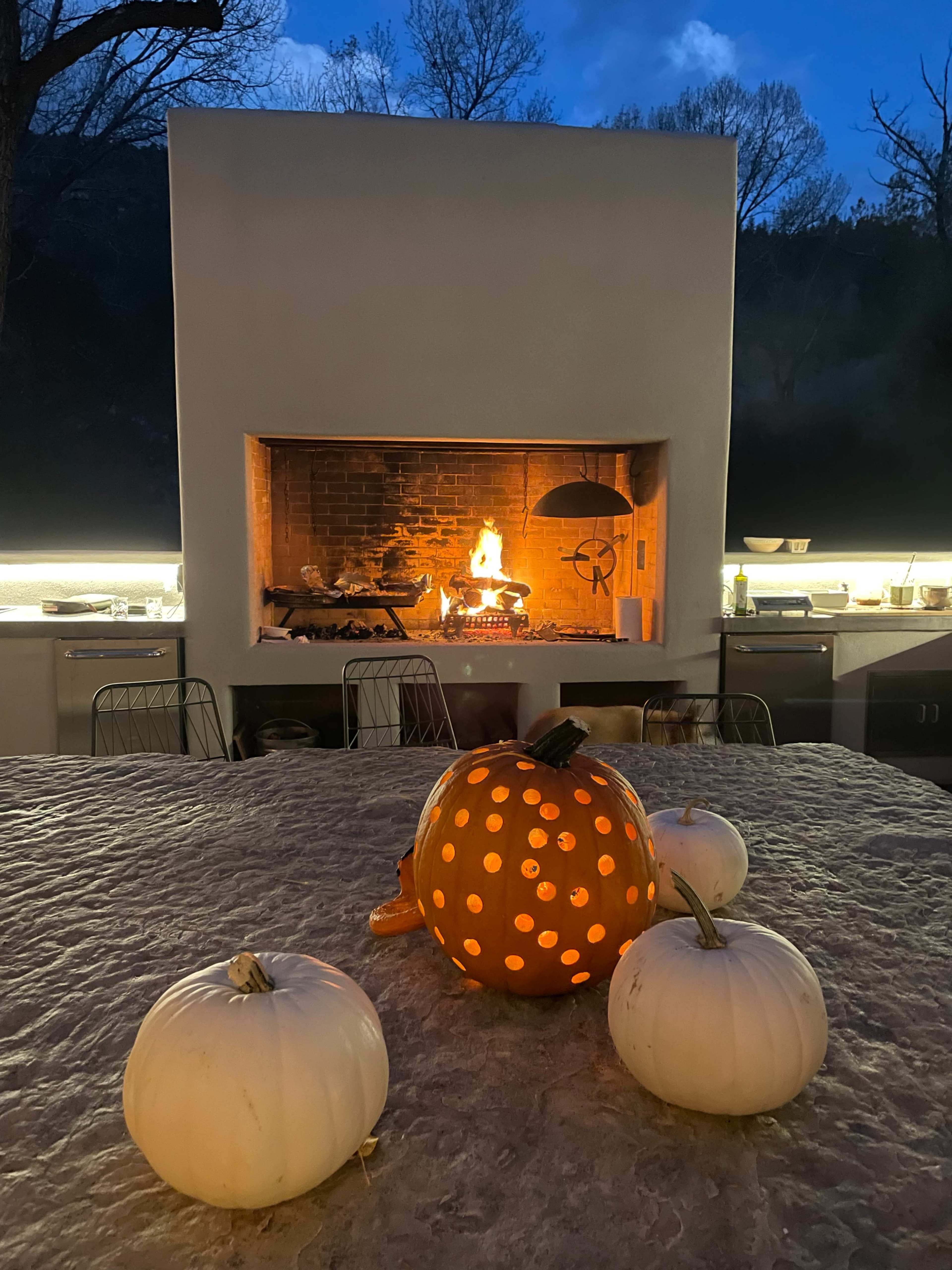 An outdoor kitchen is illuminated at dusk, featuring a lit fireplace and decorated with carved and uncarved pumpkins on a stone countertop.
