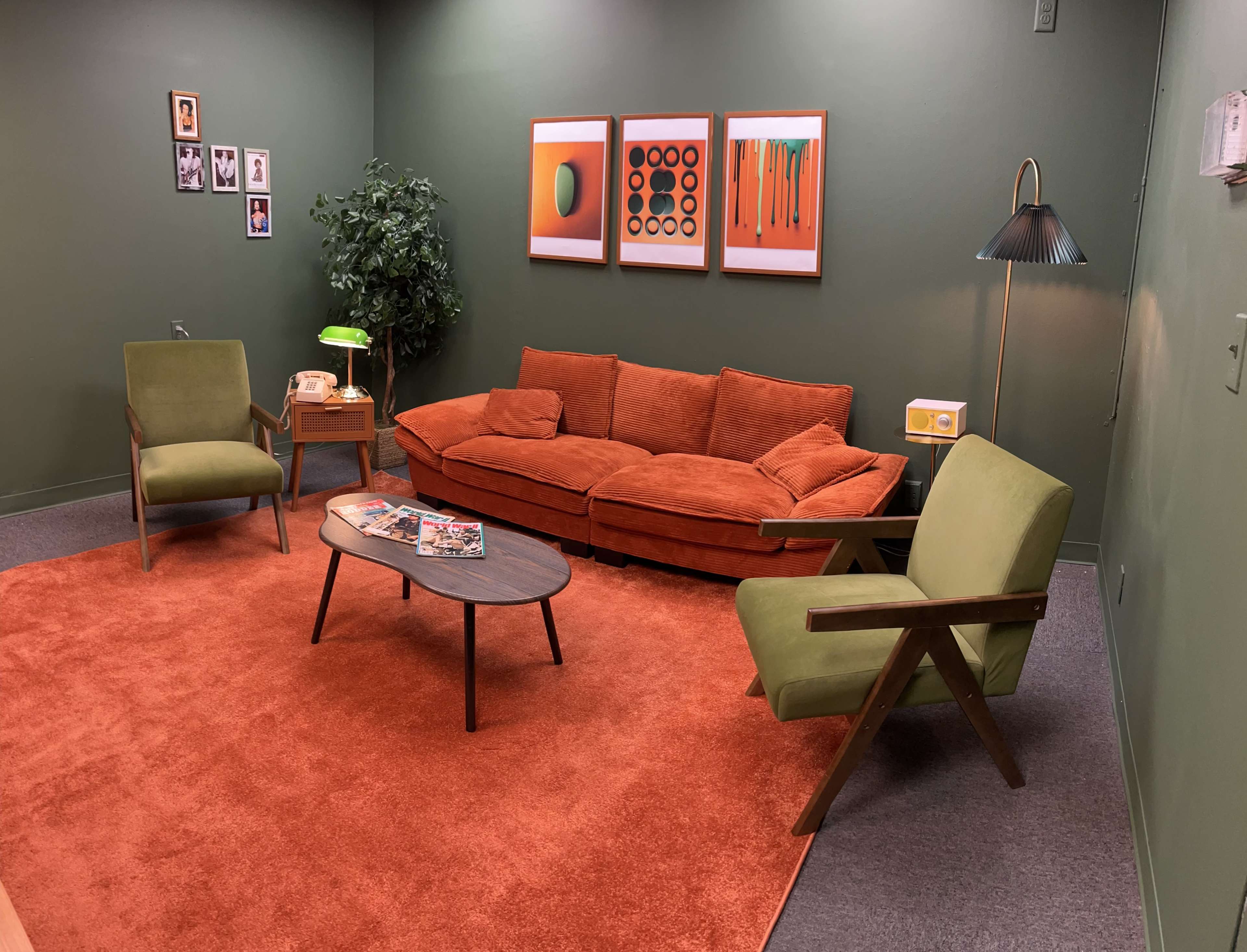 The image shows a retro-styled living room featuring an orange sectional sofa, green chairs, a circular coffee table, and colorful wall art against a green backdrop.