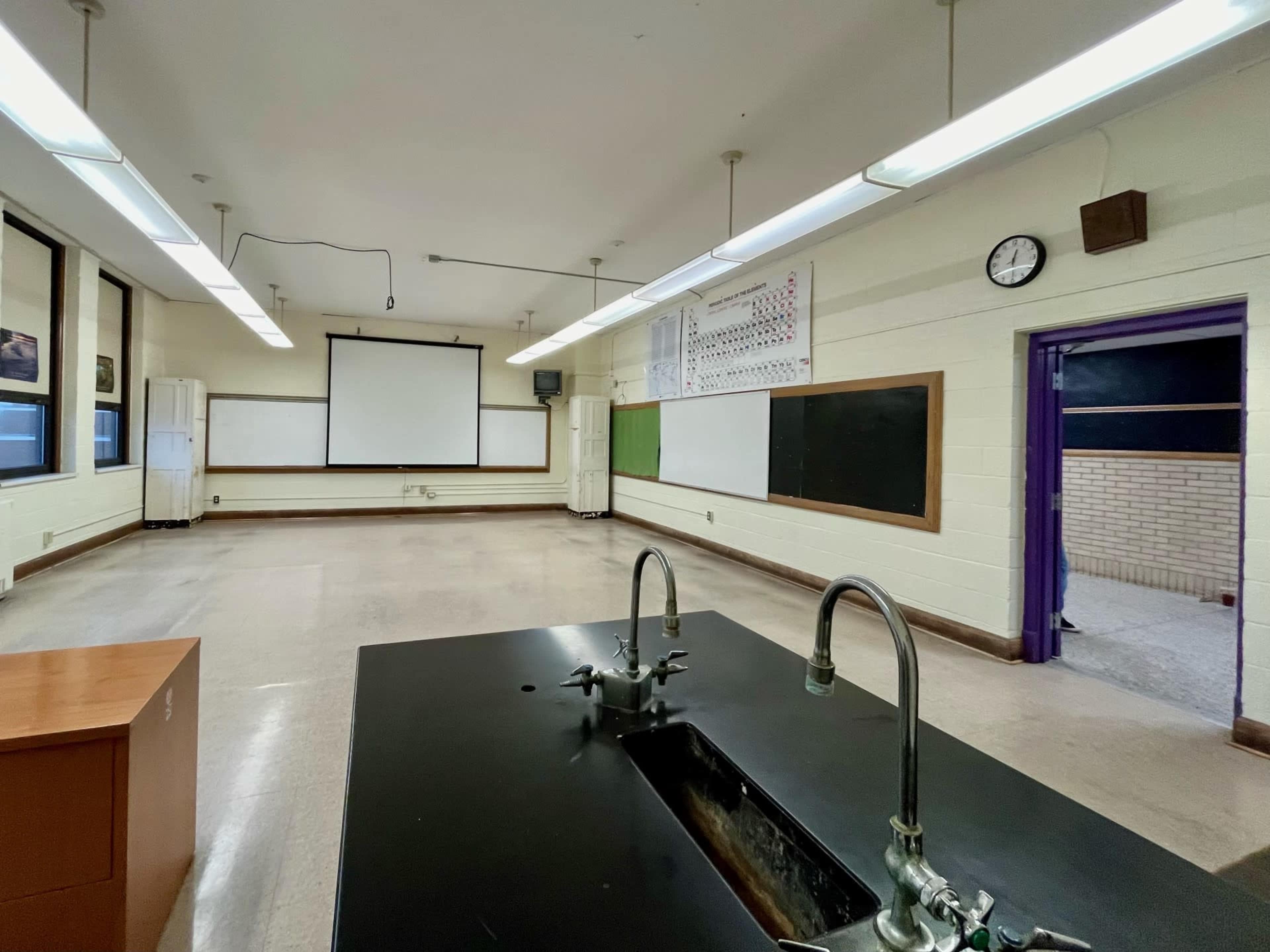 The image shows an empty classroom with a large black countertop in the foreground, a projector screen on the wall, and chalkboards on either side.