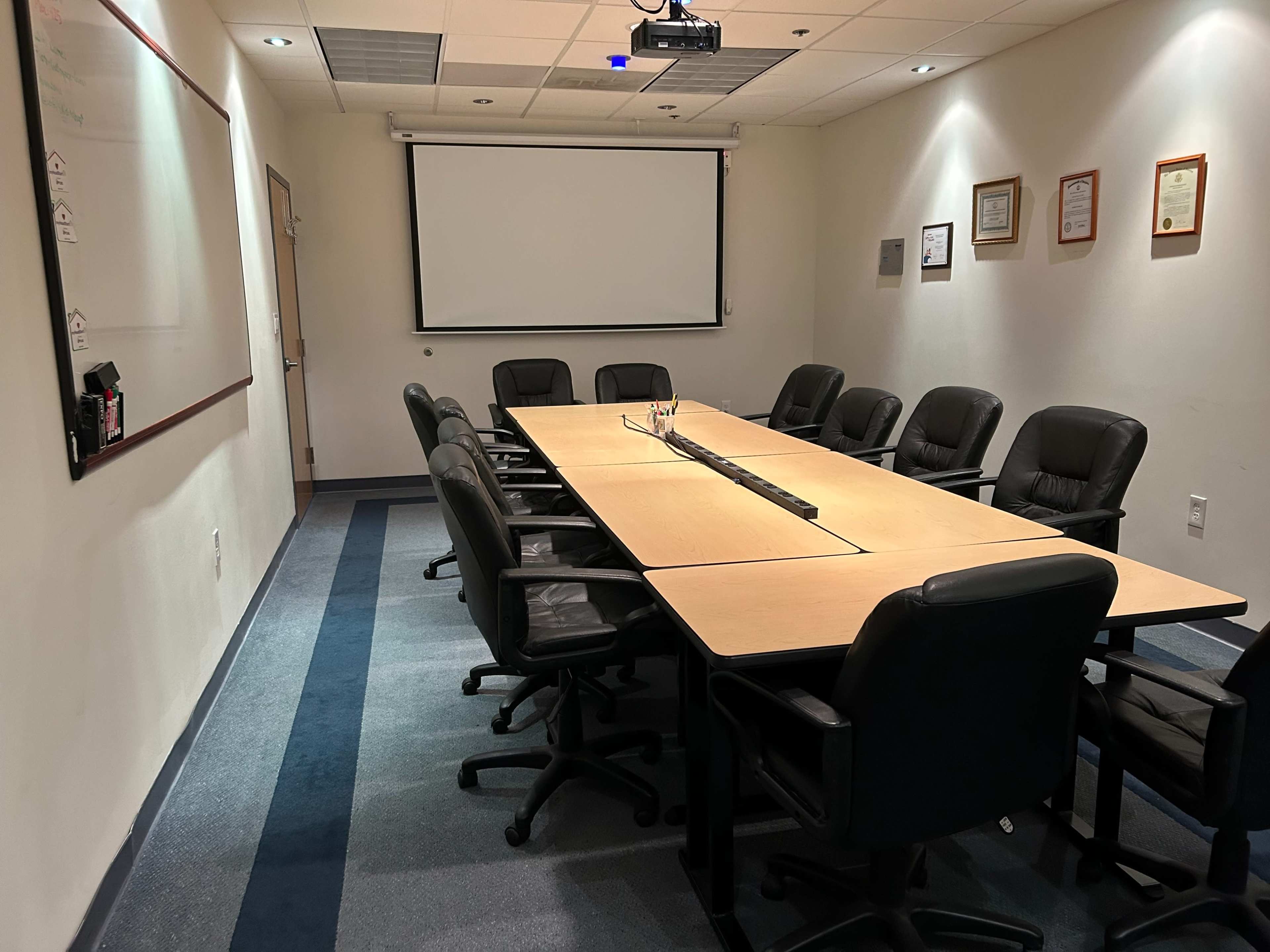 A conference room features a long rectangular table surrounded by several black chairs, with a projector screen and framed documents on the walls.