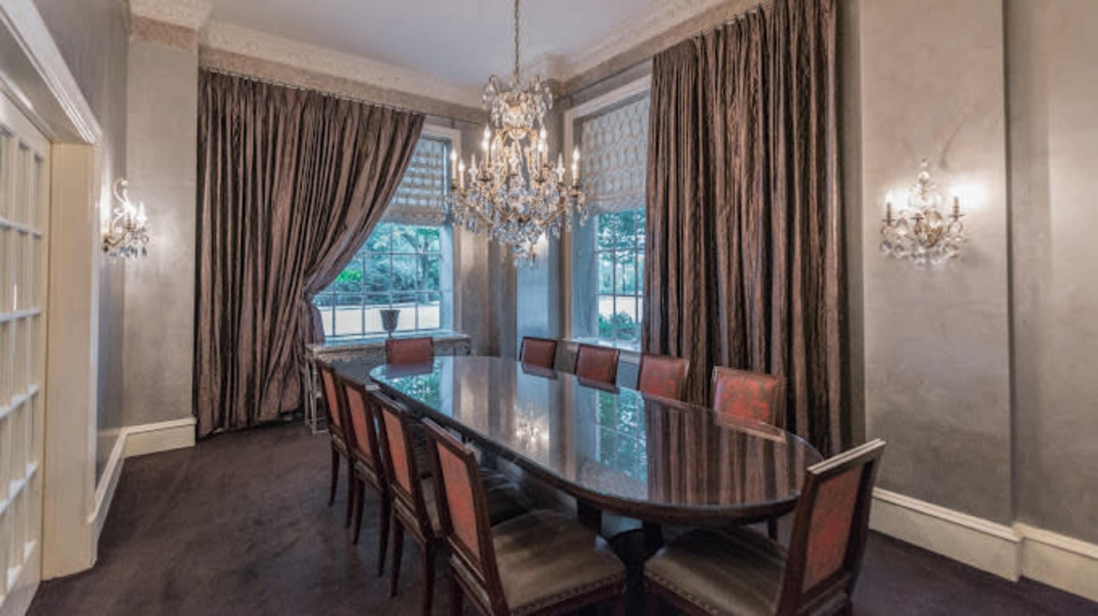 The image shows a dining room featuring a long table surrounded by upholstered chairs, with large windows and elegant drapes.