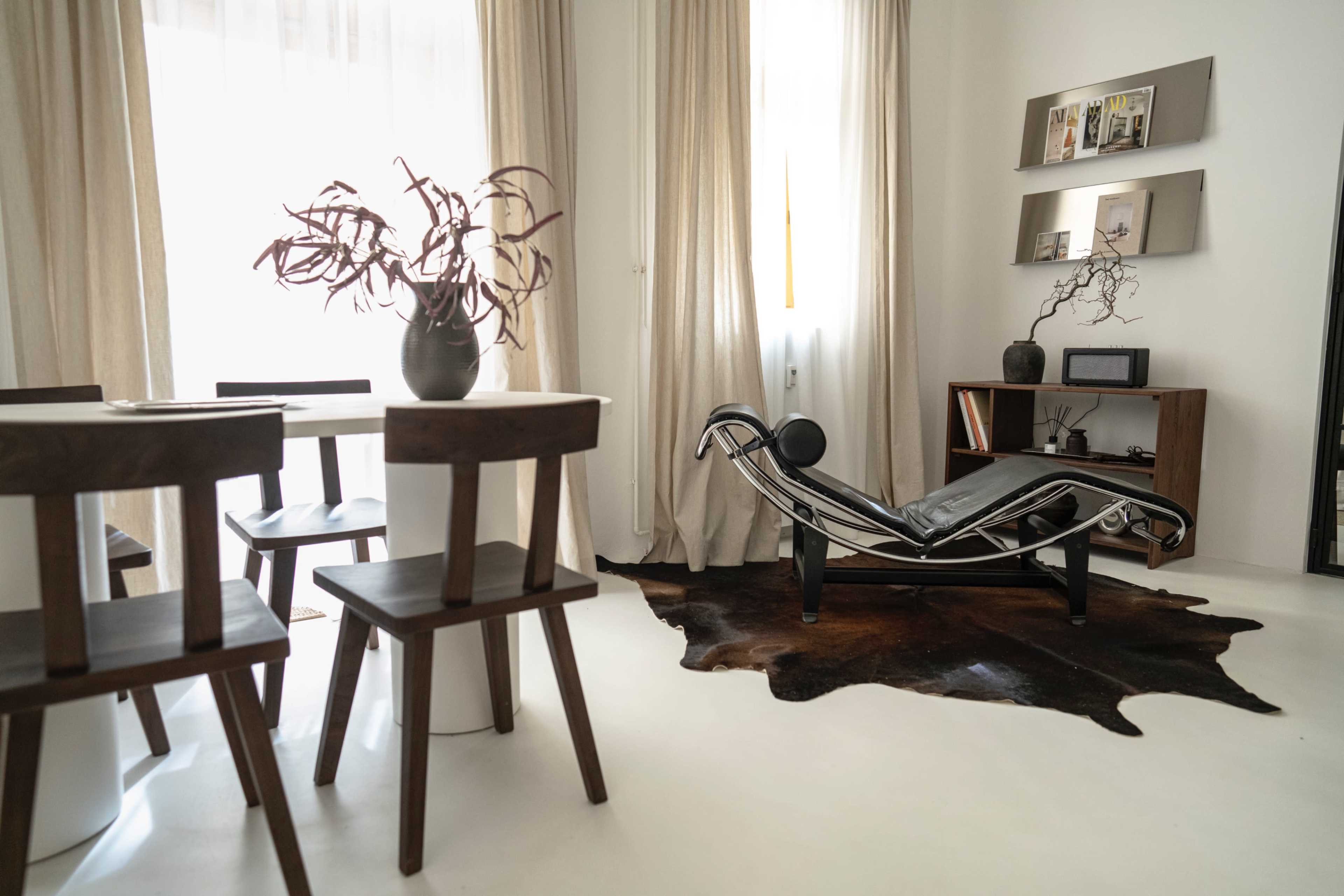 The image shows a minimalist living space featuring a dining area with wooden chairs, a lounge chair, and a stylish decorative cowhide rug on a white floor.