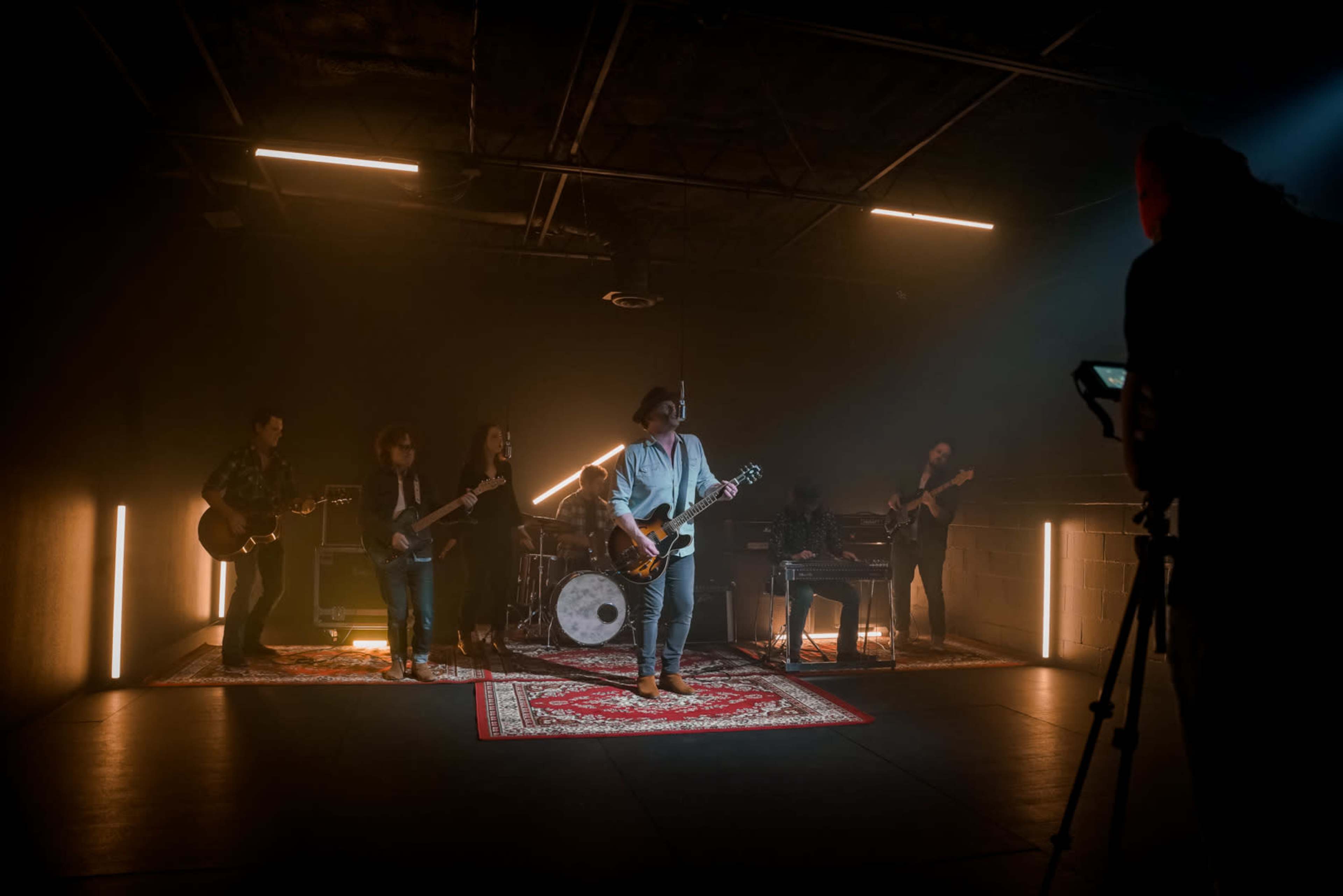 A band performs under dramatic lighting in a studio setting, with a cameraman filming from the side.