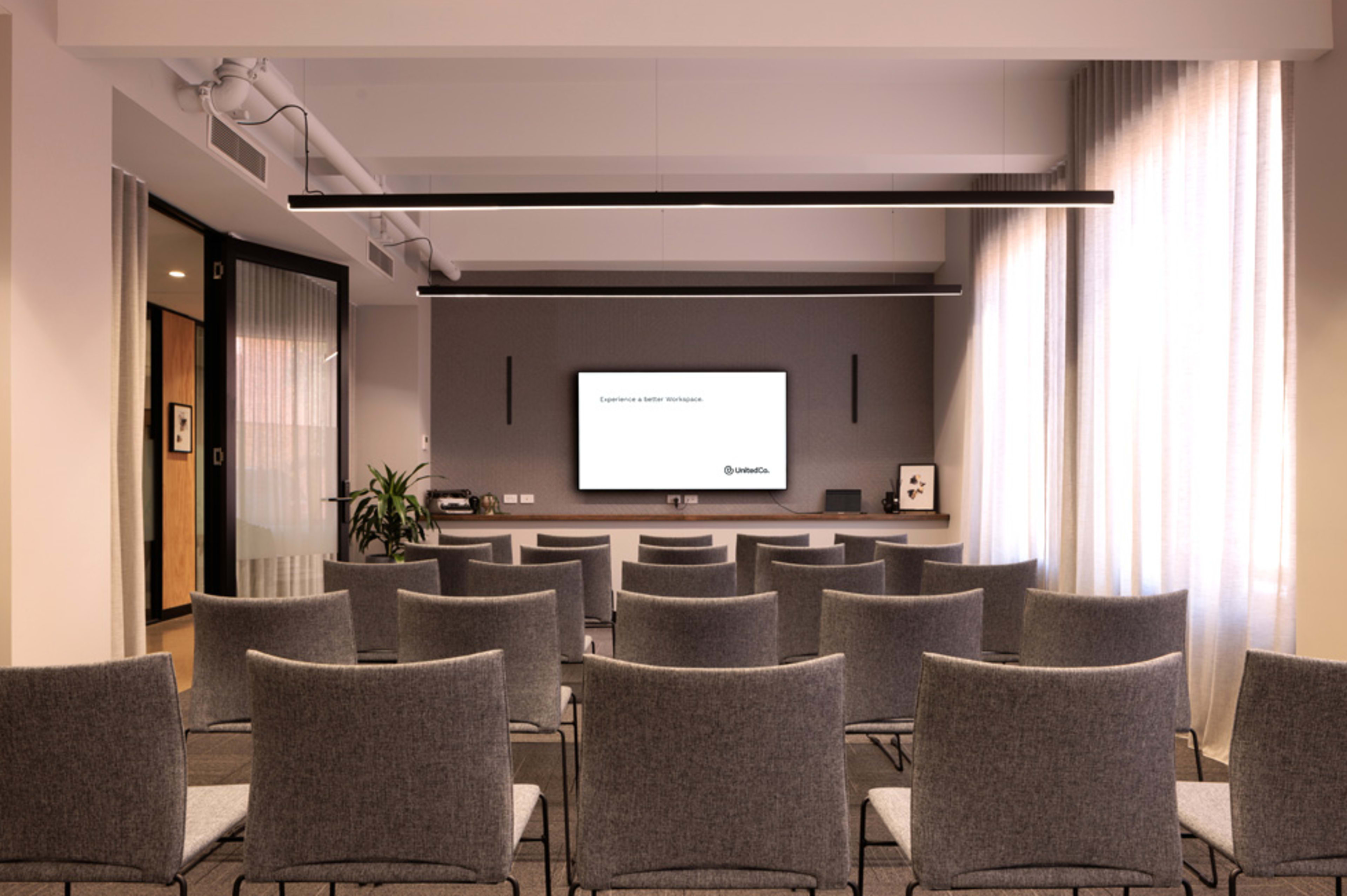 A modern conference room filled with gray chairs arranged in rows facing a mounted screen on a wall.