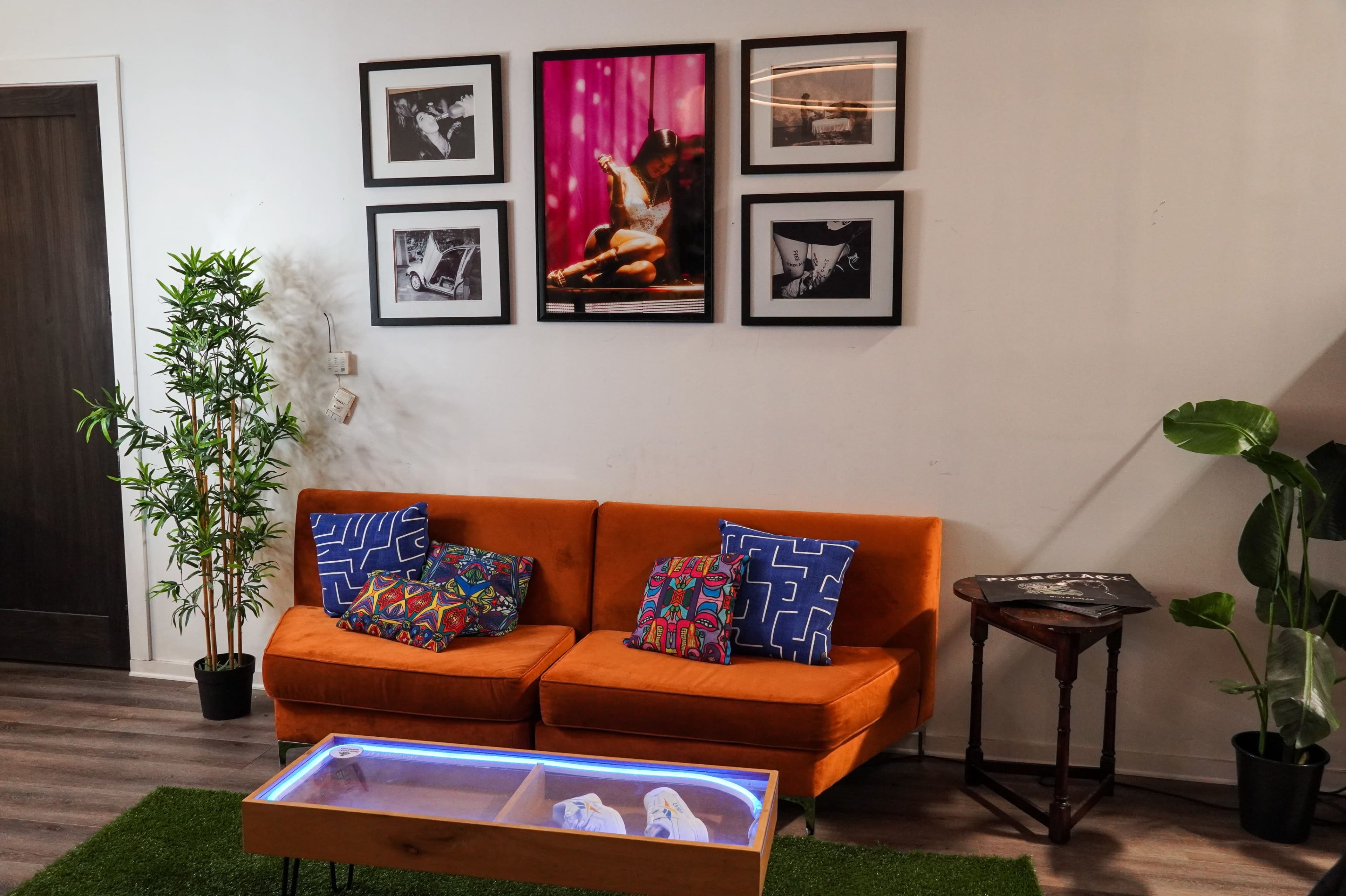 The image shows a cozy living space featuring an orange couch with decorative pillows, a glass coffee table, framed photographs on the wall, and some indoor plants.