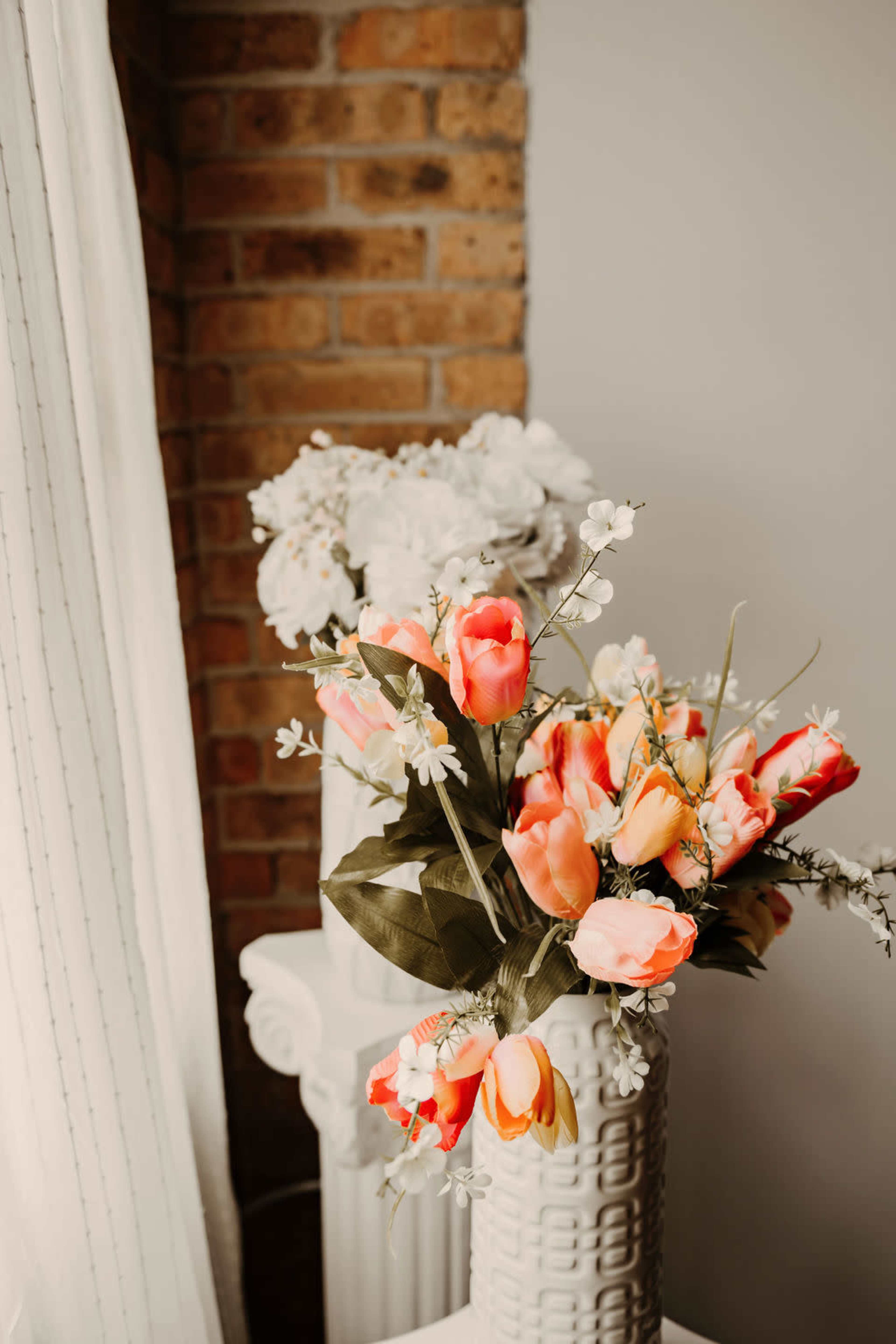 A bouquet of artificial flowers, including orange tulips and white blossoms, is arranged in a textured white vase on a decorative column, with a brick wall in the background.