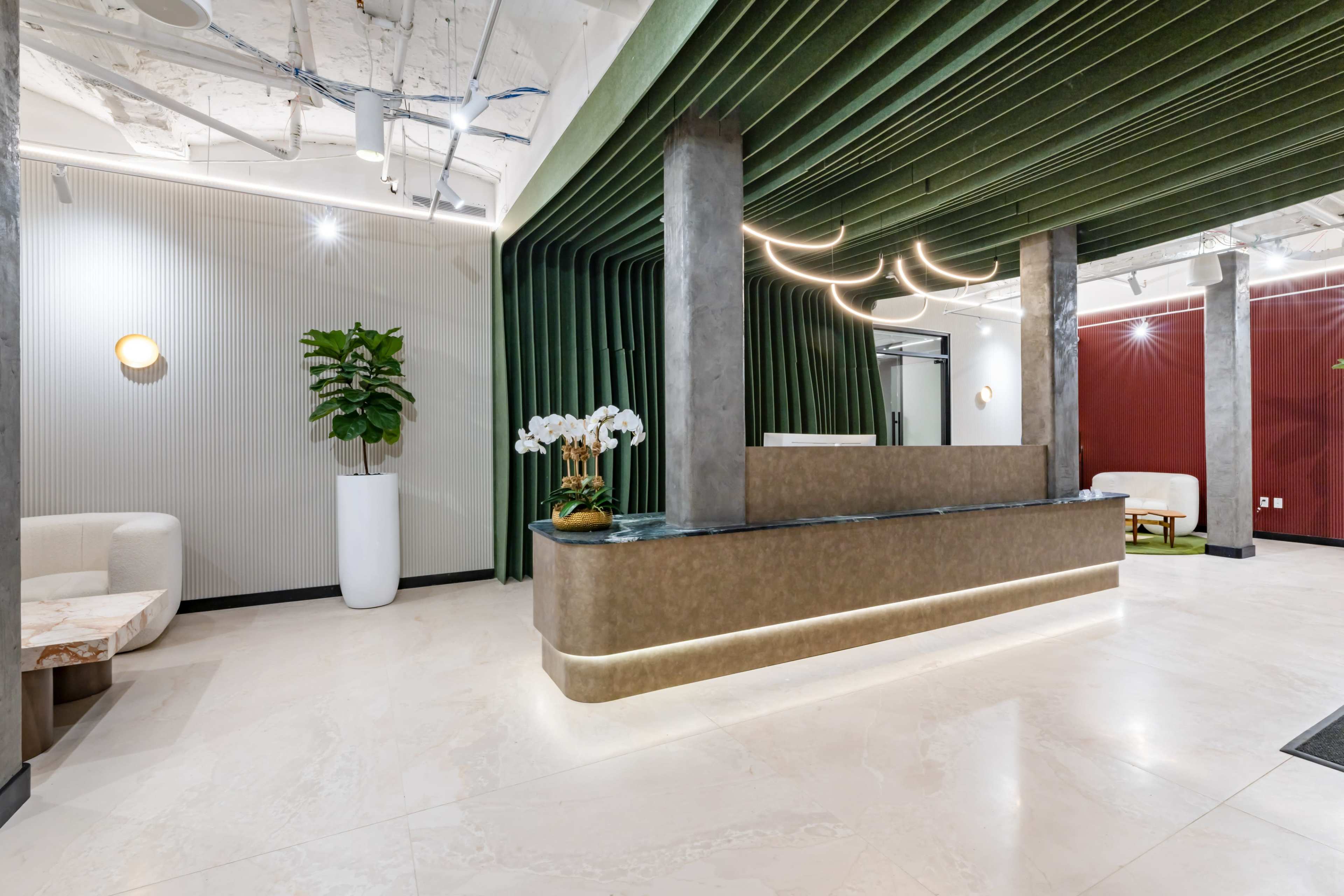 The image shows a modern reception area with a curved front desk, green fabric walls, streamlined lighting, and decorative plants.