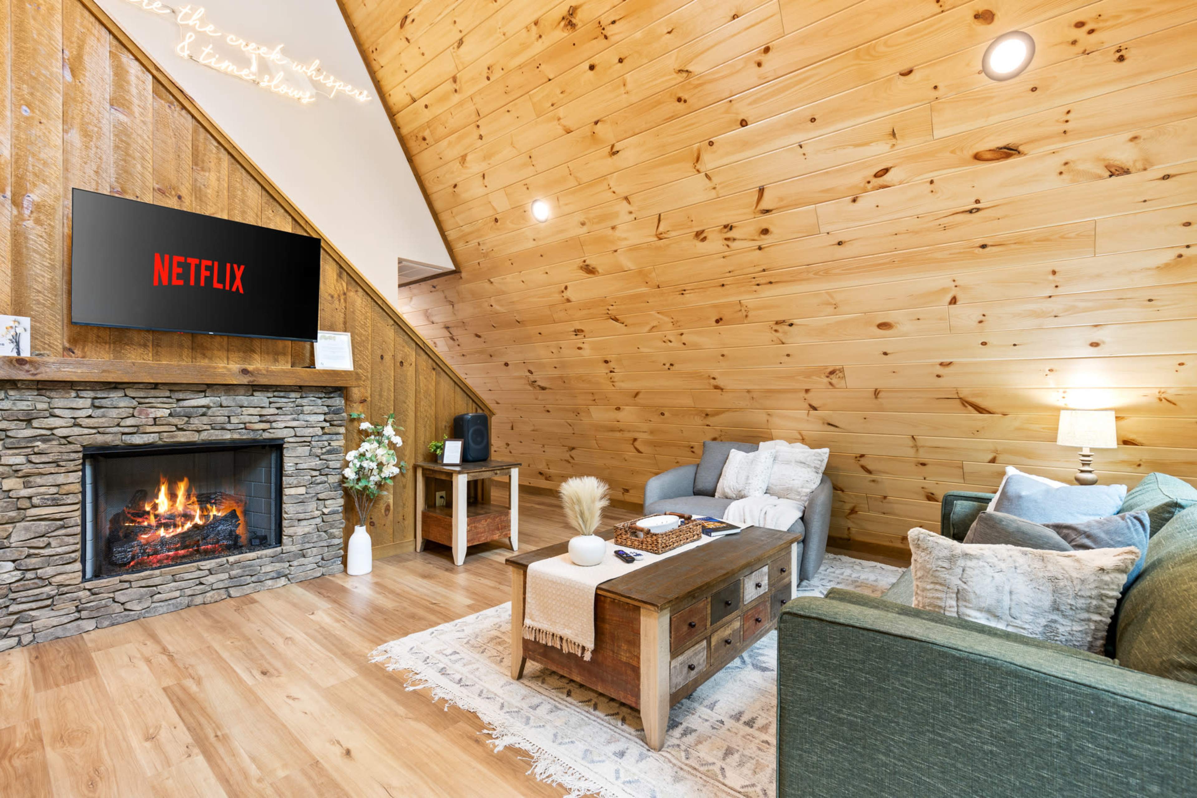 The image shows a cozy living room with wooden walls, a stone fireplace, comfortable seating, and a television displaying the Netflix logo.