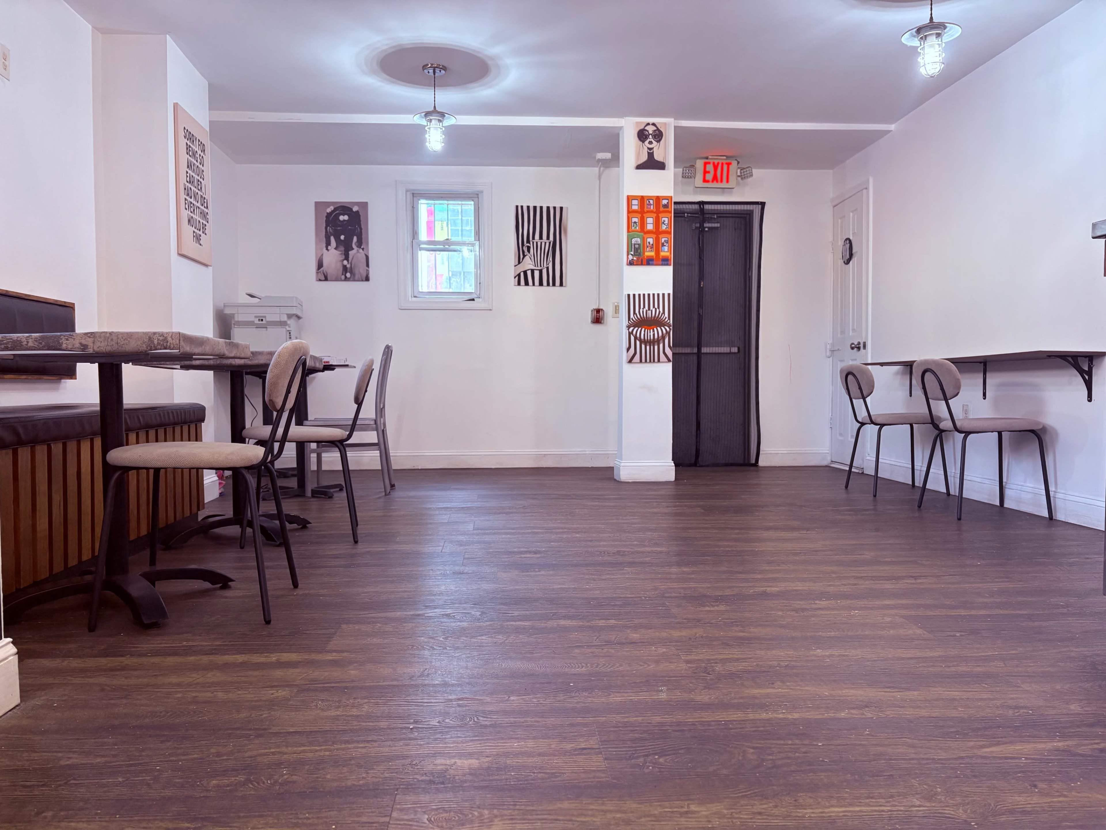 The image shows a spacious, empty room with wooden flooring, several chairs and tables, and various artworks on the walls.