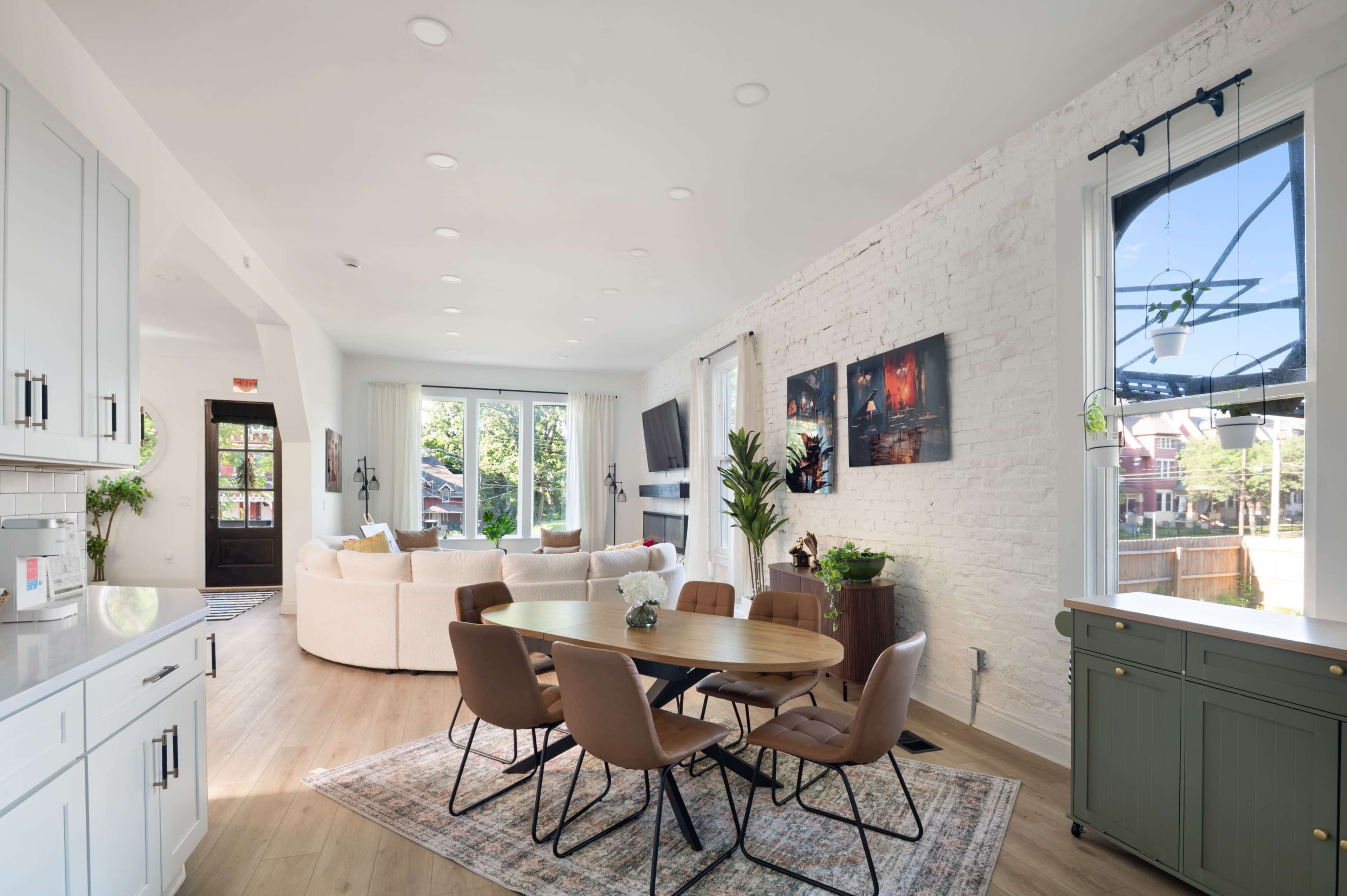 The image shows a modern dining area with a round wooden table surrounded by brown chairs, near a large window that lets in natural light, with a living space visible in the background.