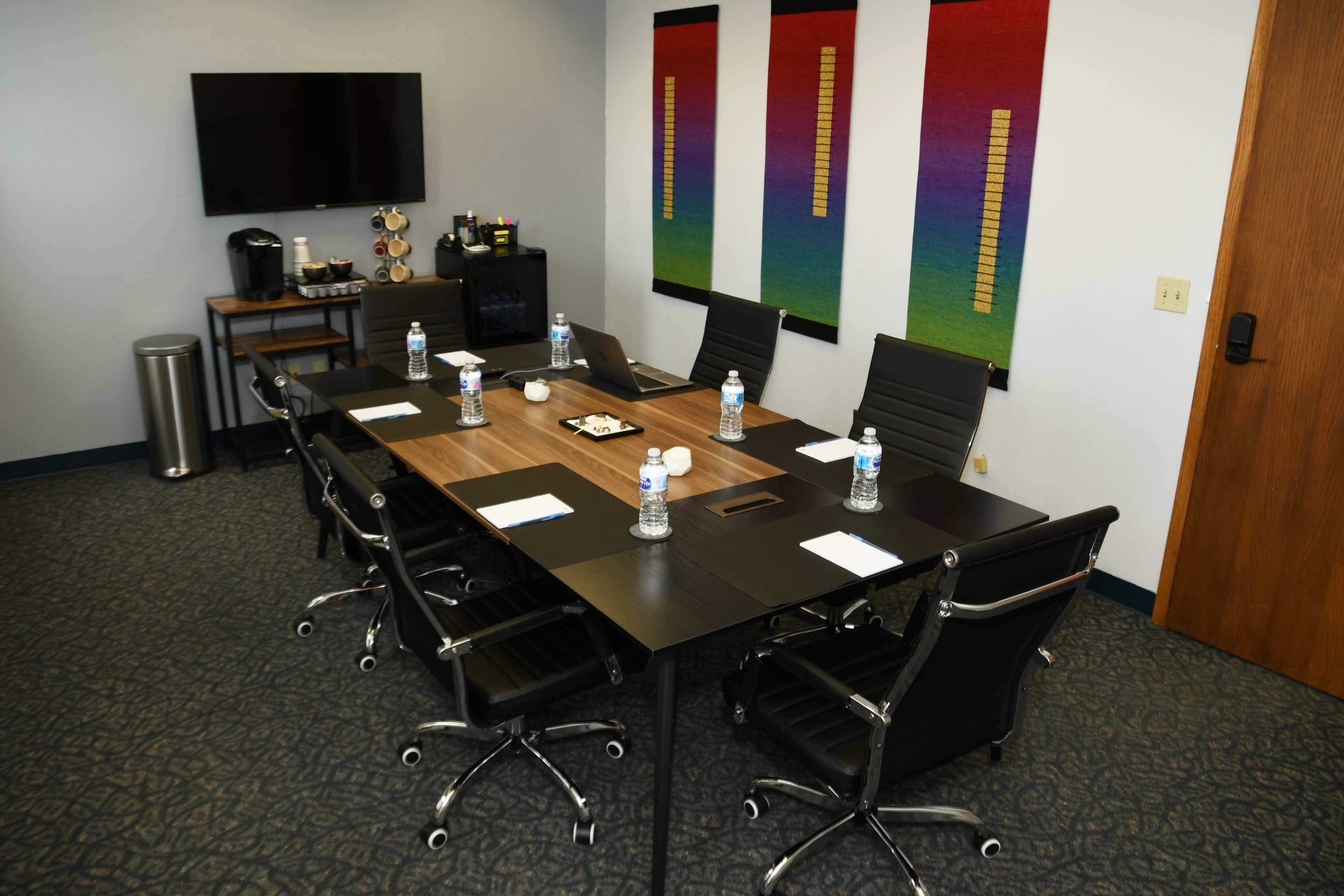 The image depicts a modern conference room featuring a rectangular table with black chairs, a television, and colorful vertical artwork on the walls.