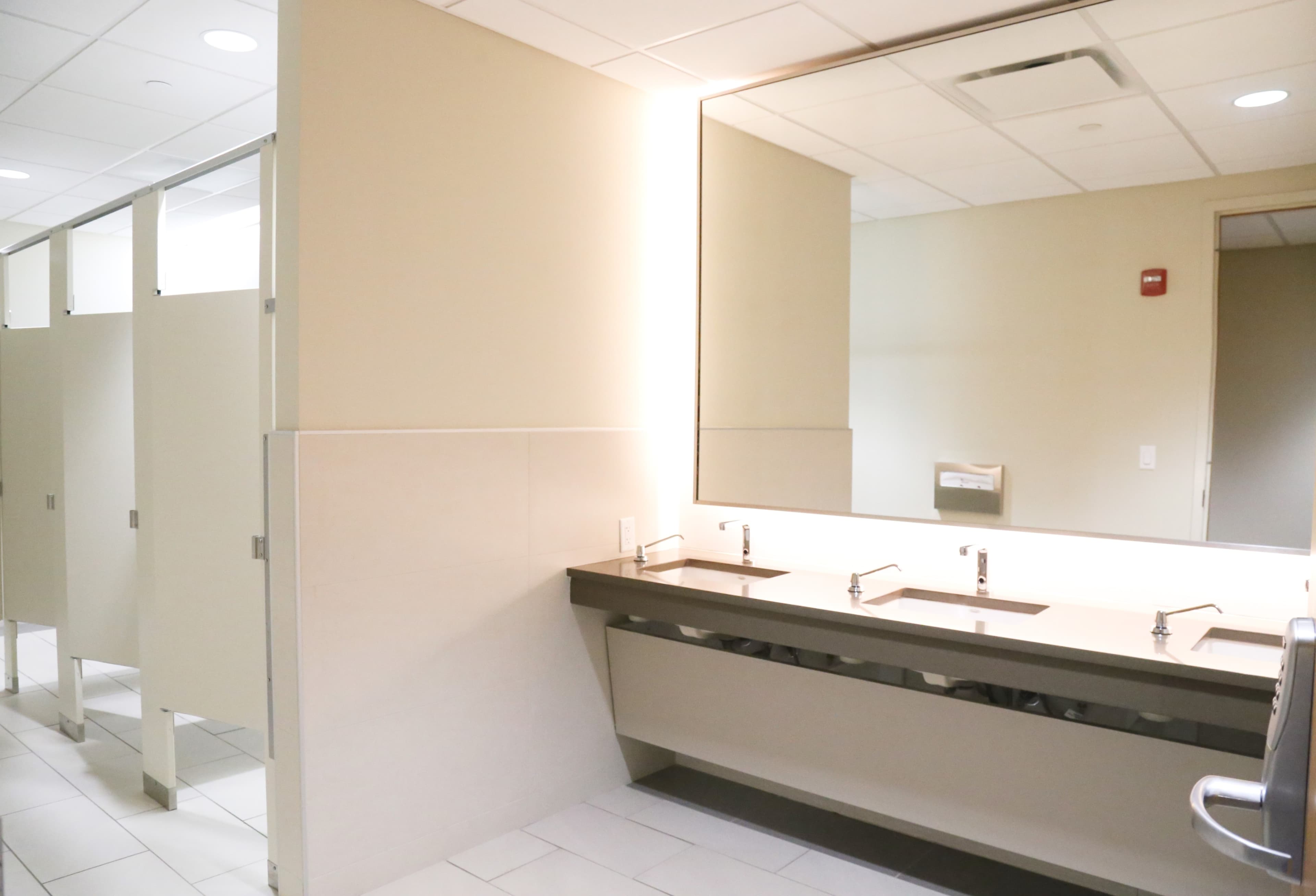 A restroom features a long sink area with two faucets and a row of privacy stalls on one side.