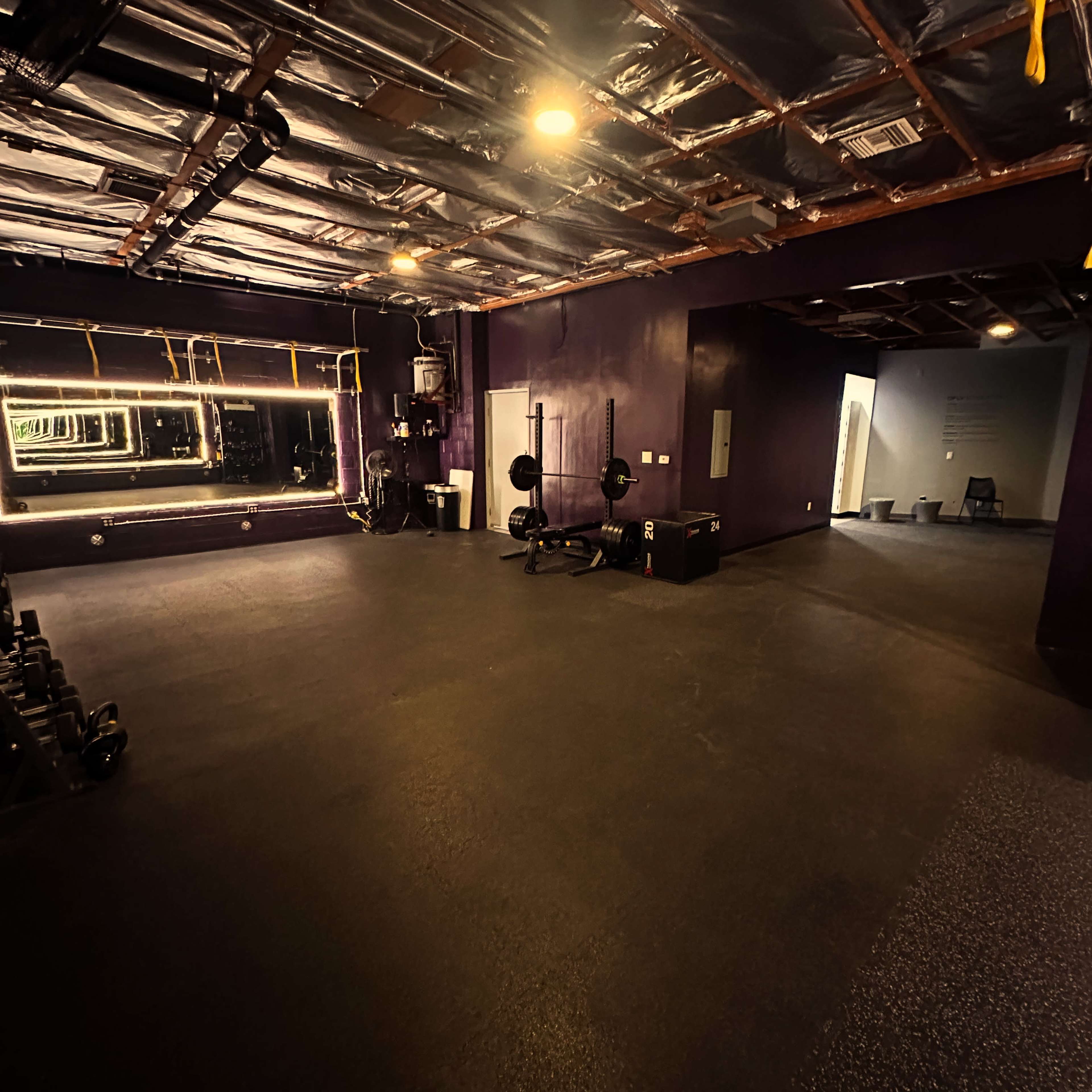 The image shows an empty gym space with purple walls, a mirror, dumbbells, a weight bench, and a clear area leading to another room.