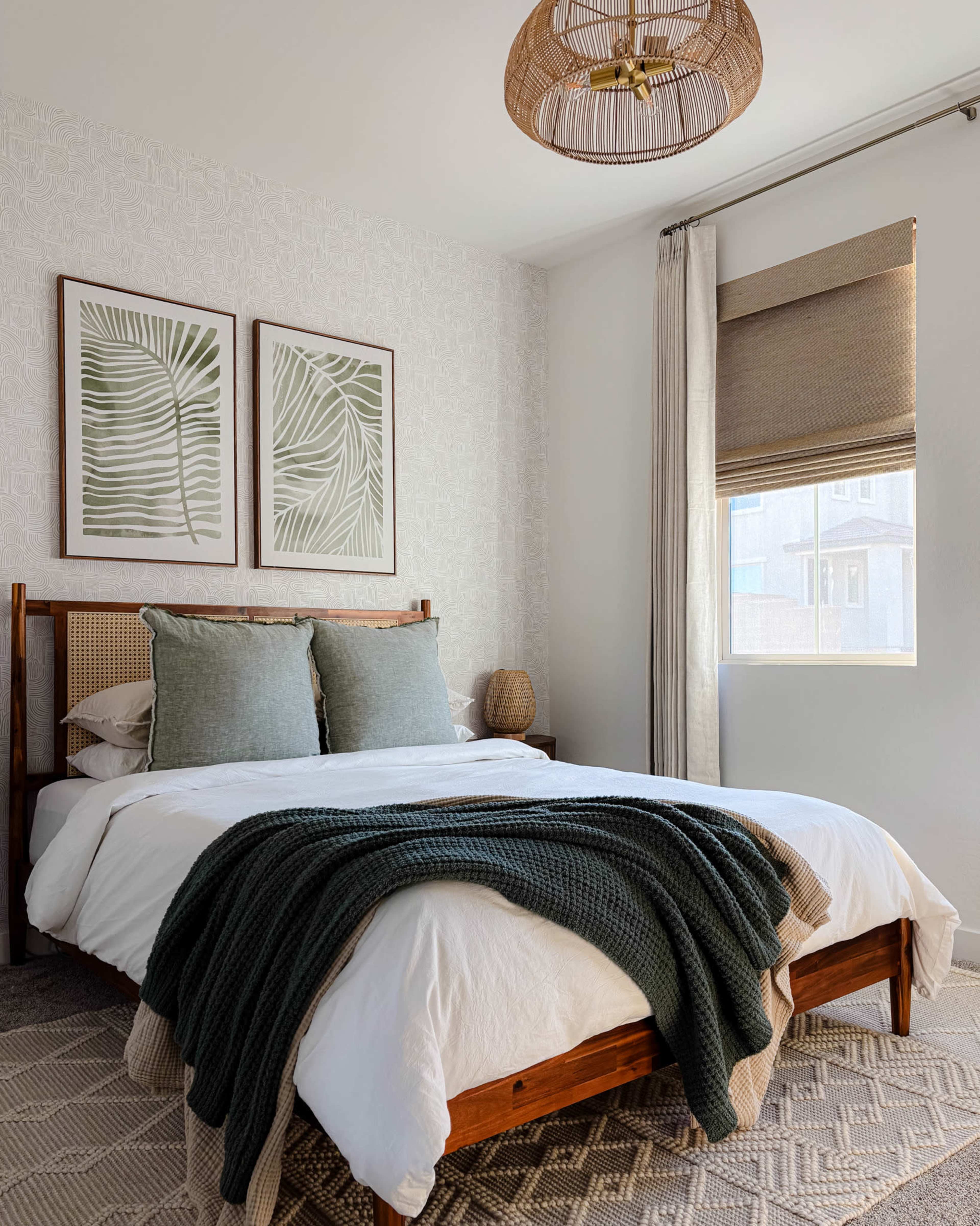 The image shows a bright bedroom featuring a wooden bed with a white comforter, green accent pillows, and two framed leaf prints on the wall.