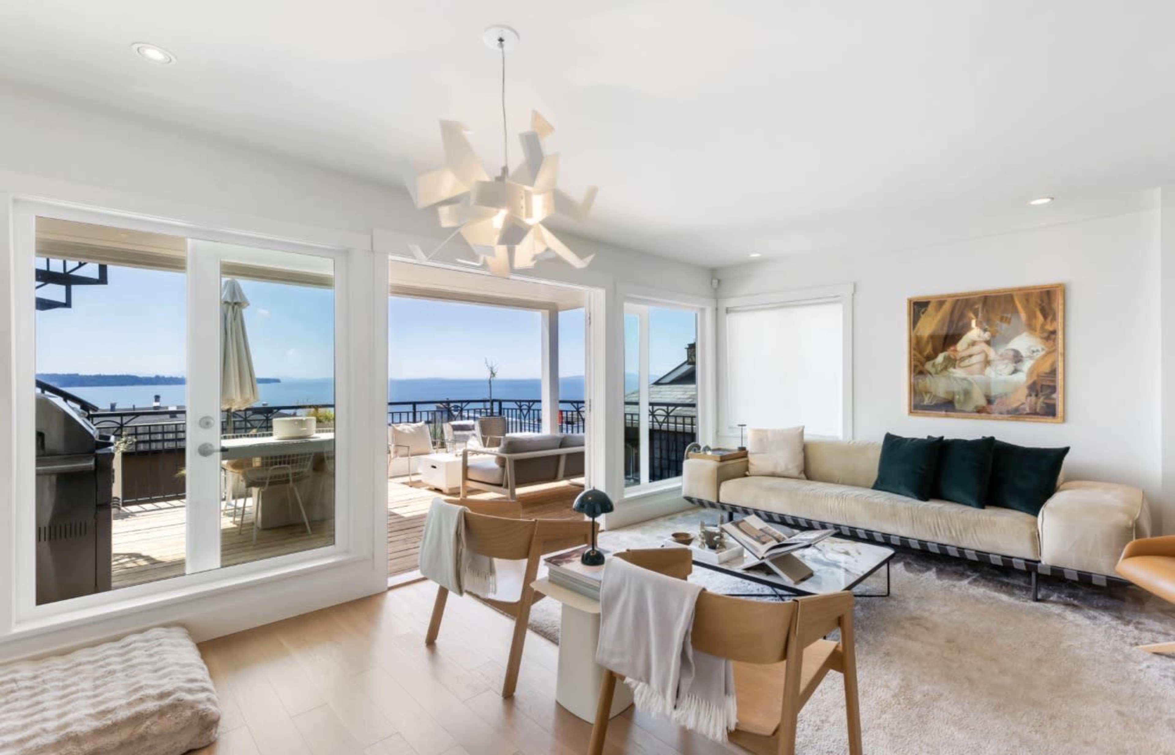 The image shows a modern living room with large windows overlooking a body of water, featuring a light-colored sofa, wooden chairs, and a coffee table.