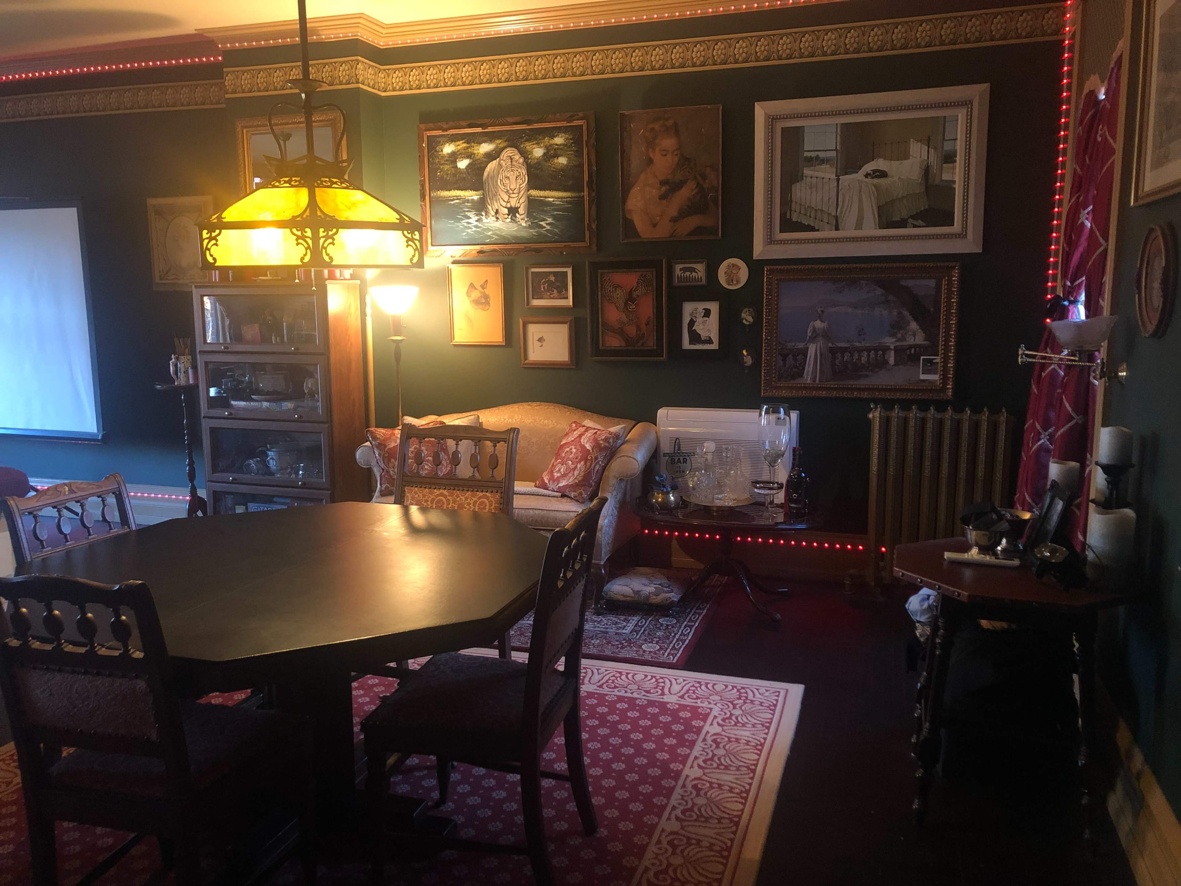 A vintage-style room with a dining table, upholstered sofa, and various framed artworks on the walls, illuminated by a decorative lamp.