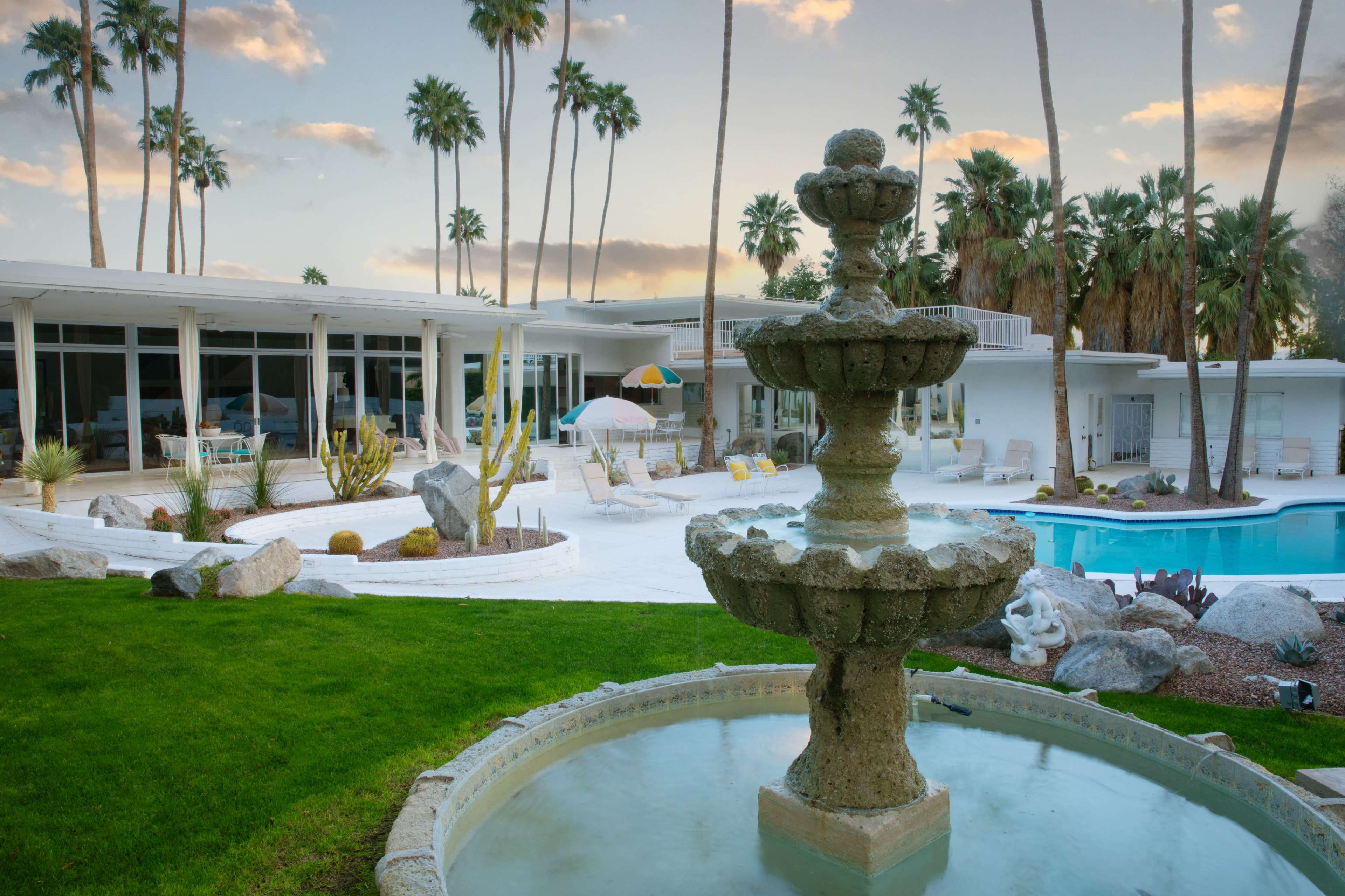 The image shows a modern backyard with a fountain in the foreground, a swimming pool, lounge chairs, and palm trees.