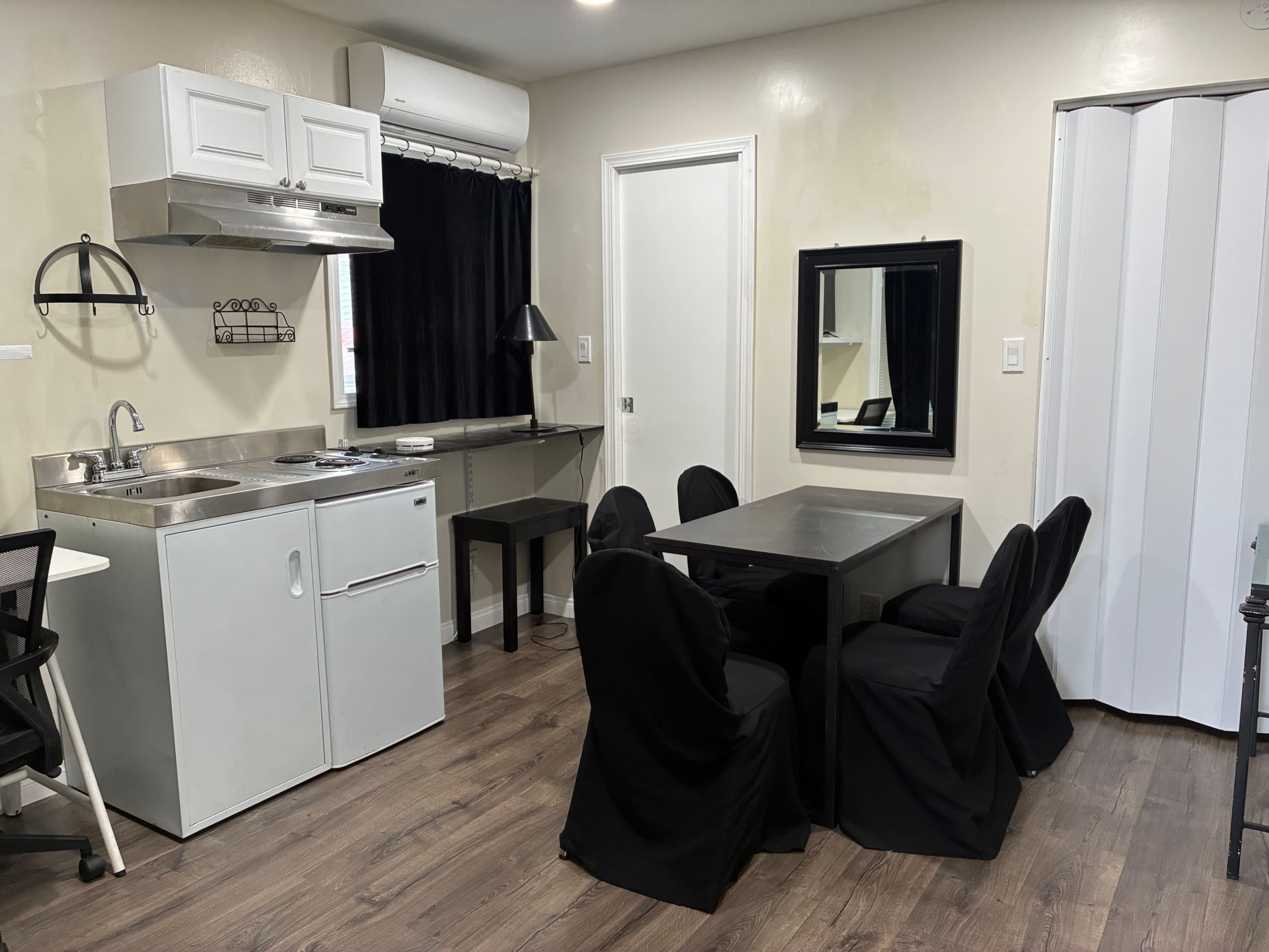 The image shows a small kitchen and dining area with a table surrounded by chairs, a kitchenette with a sink and stove, and a mirror on the wall.