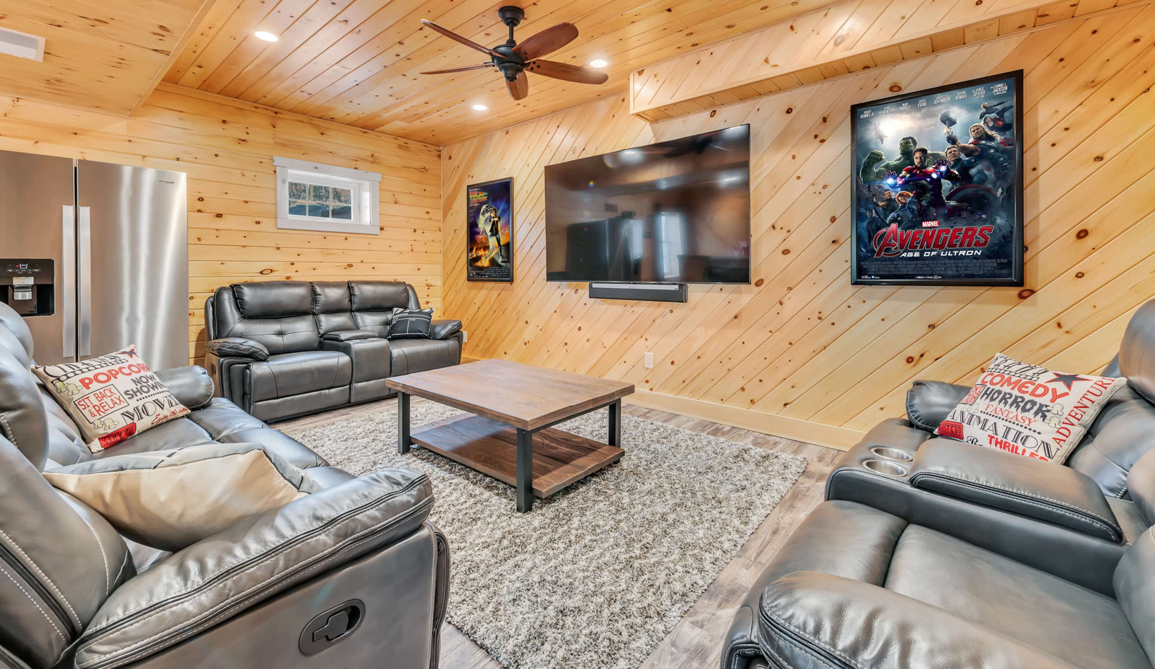 A cozy home theater features leather recliners, a coffee table, and a large wall-mounted television, with wooden paneling and movie posters decorating the space.